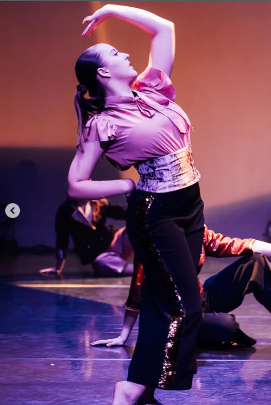 Woman dancing on stage in beige top and black sequined trousers, one arm raised overhead, under purple and pink stage lighting.