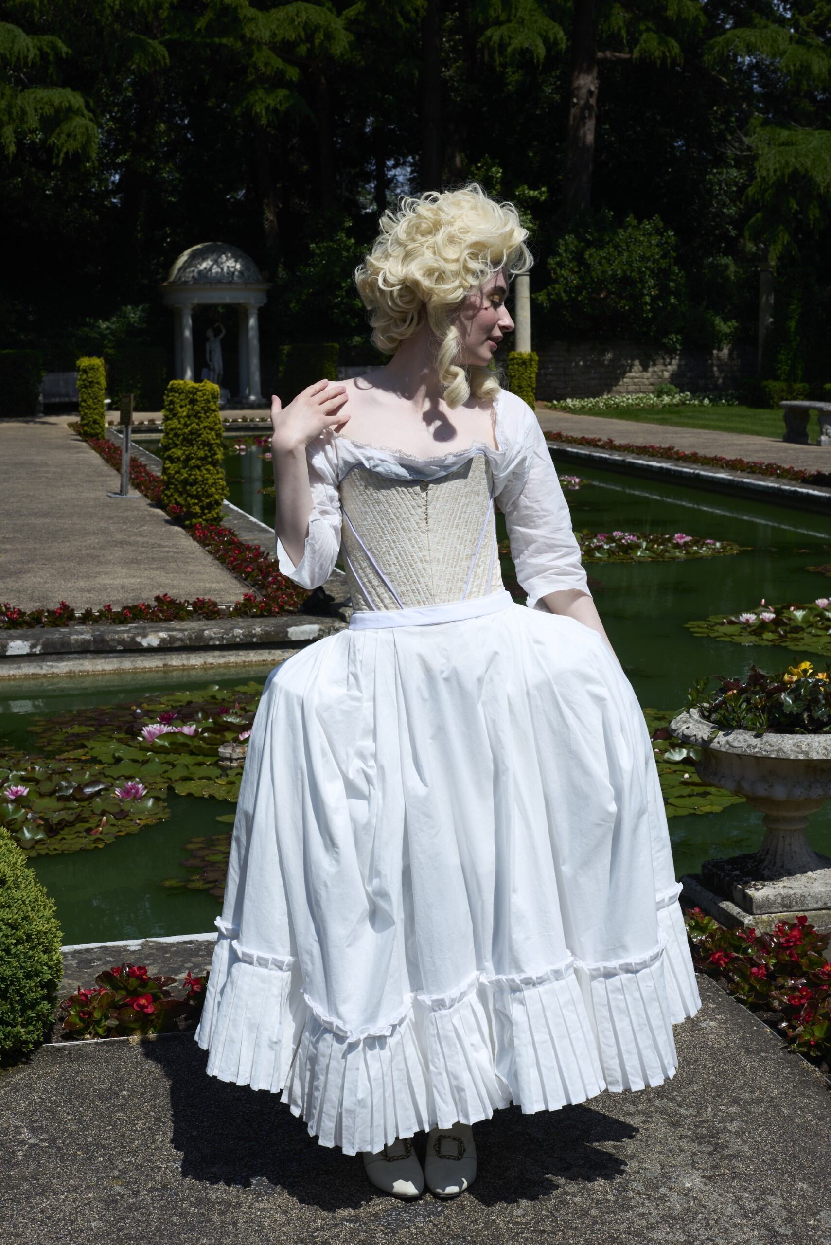 Woman in white 18th-century costume with powdered blonde wig standing in formal gardens with hedgerows and pavilion.