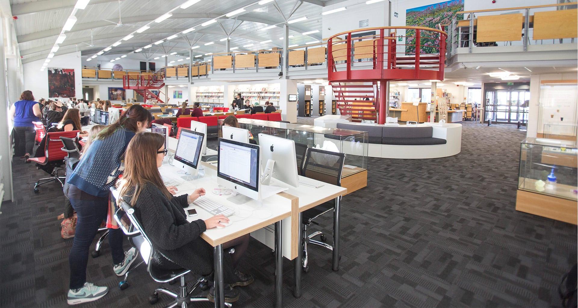 Students working at computers in a library. A set of stairs leads up to a second landing with a balcony running along.