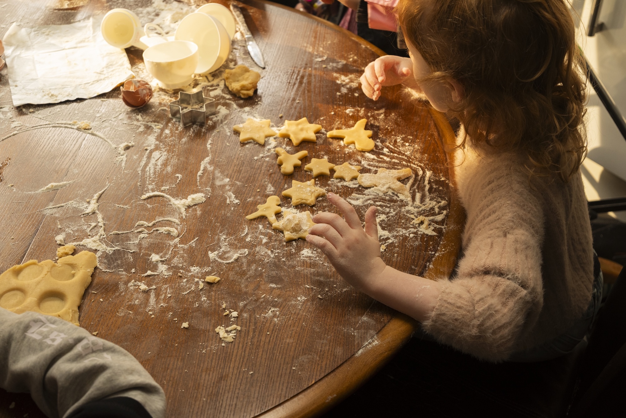 Child's hands kneading yellow dough on flour-dusted wooden surface with cookie cutters and baking ingredients nearby.