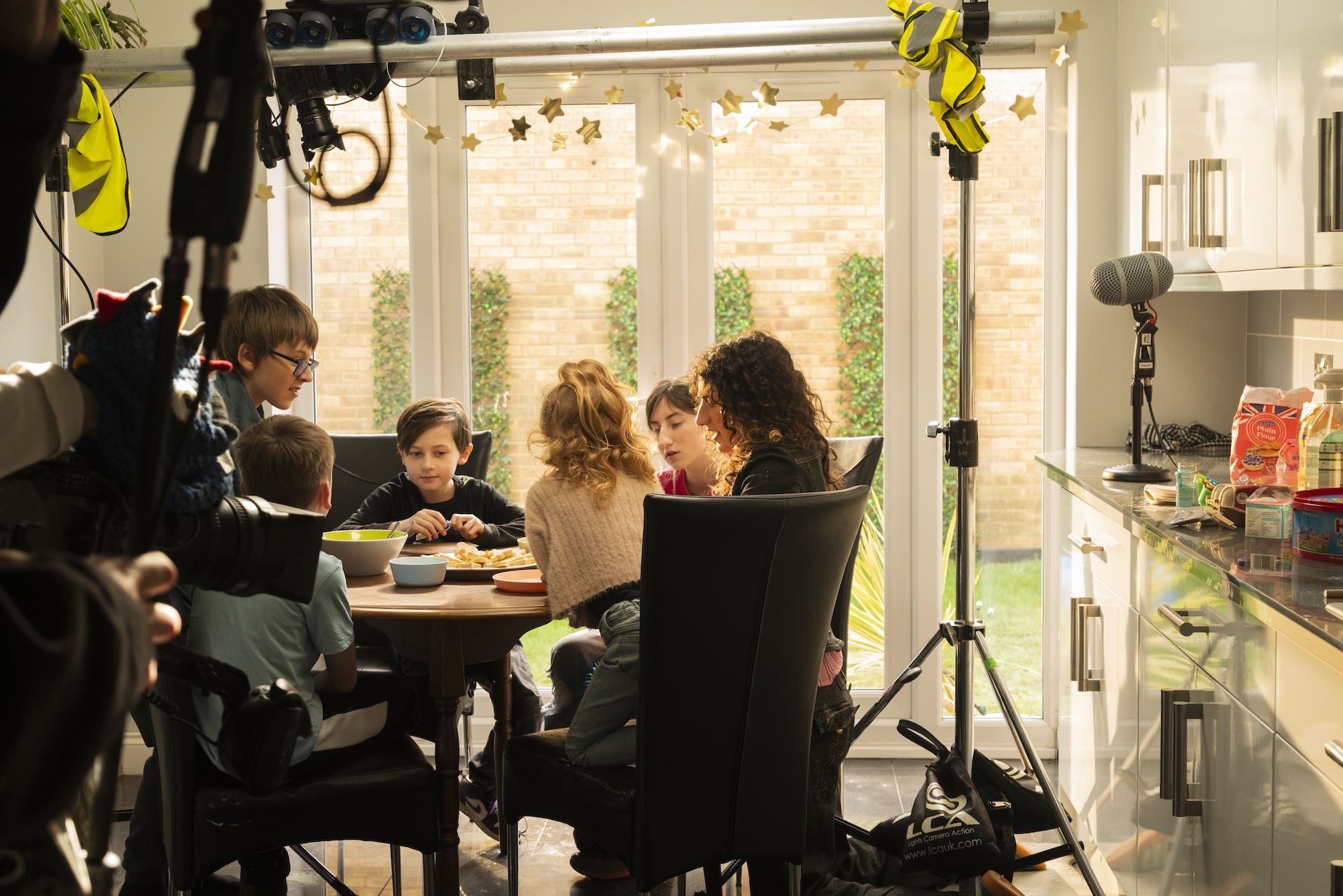 Film crew recording family of five seated round kitchen table with lighting equipment and cameras in bright modern kitchen.