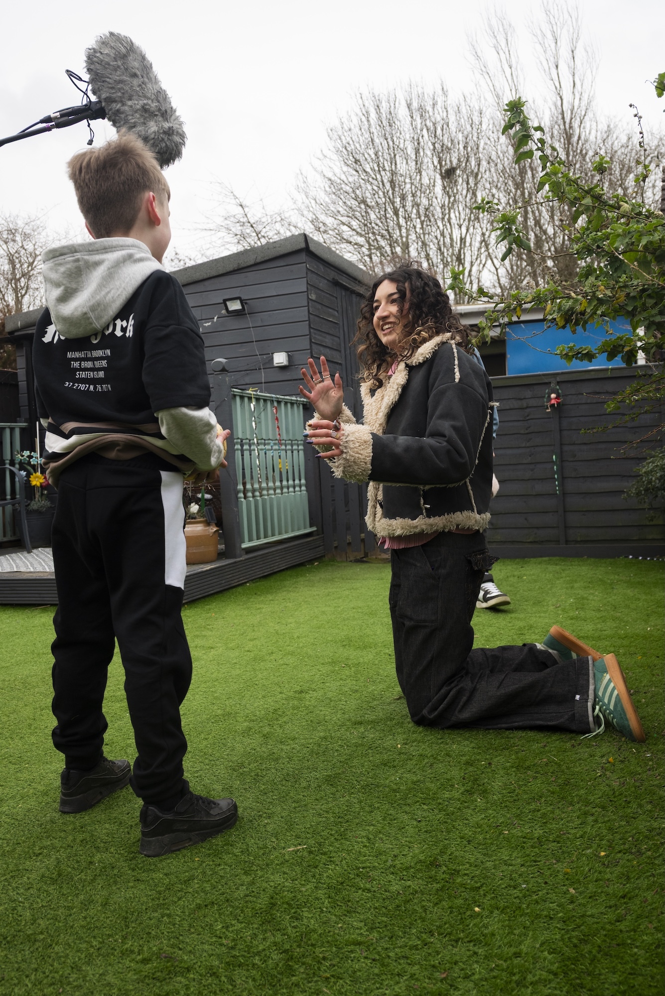 Camera operator films woman kneeling on artificial grass in back garden with grey shed and bare trees in background.