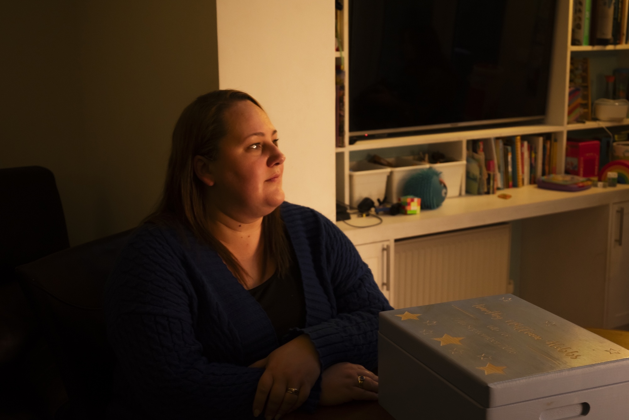 Woman in dark blue top sits at white desk, looking left. Warm lighting illuminates her face. White shelving with books visible behind.