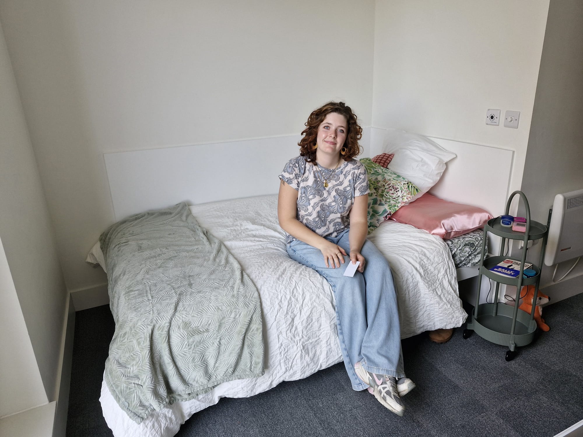 BA (Hons) Acting student R Hudson sits on a neatly made bed in a simple bedroom. The bed is covered with a white duvet and a green throw, while a small round table beside the bed holds a few items. The room has light-coloured walls and grey carpet.