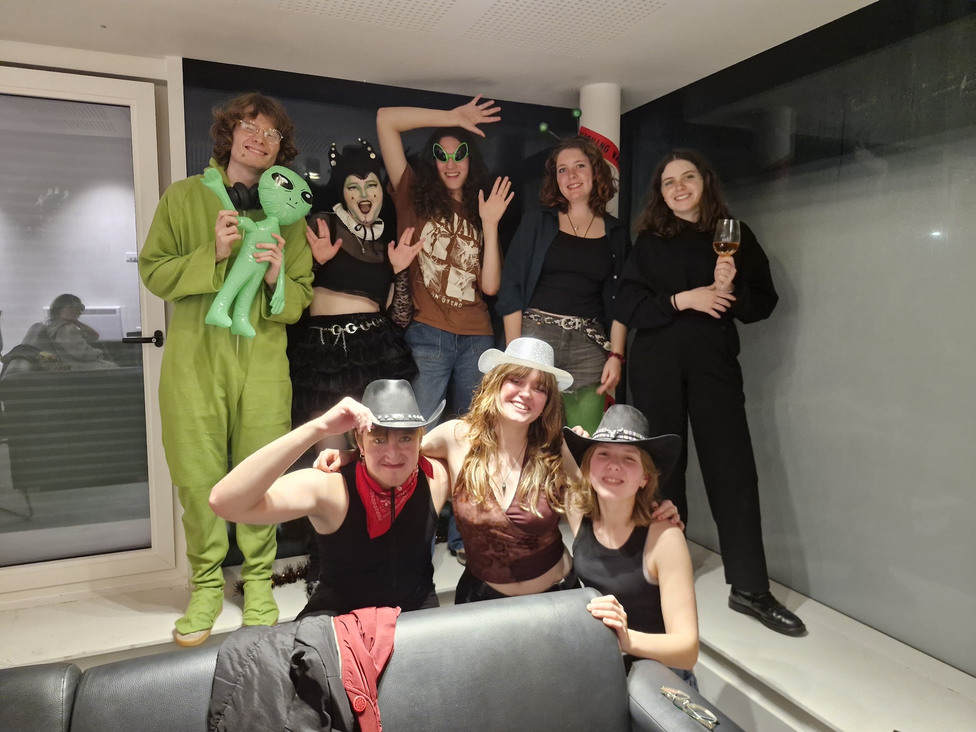 A group of eight Arts University Bournemouth students in a living room, some wearing costumes, including an alien suit, witch hat, and cowboy hats. Two people are seated in front, while others stand behind them, smiling and posing for the camera.