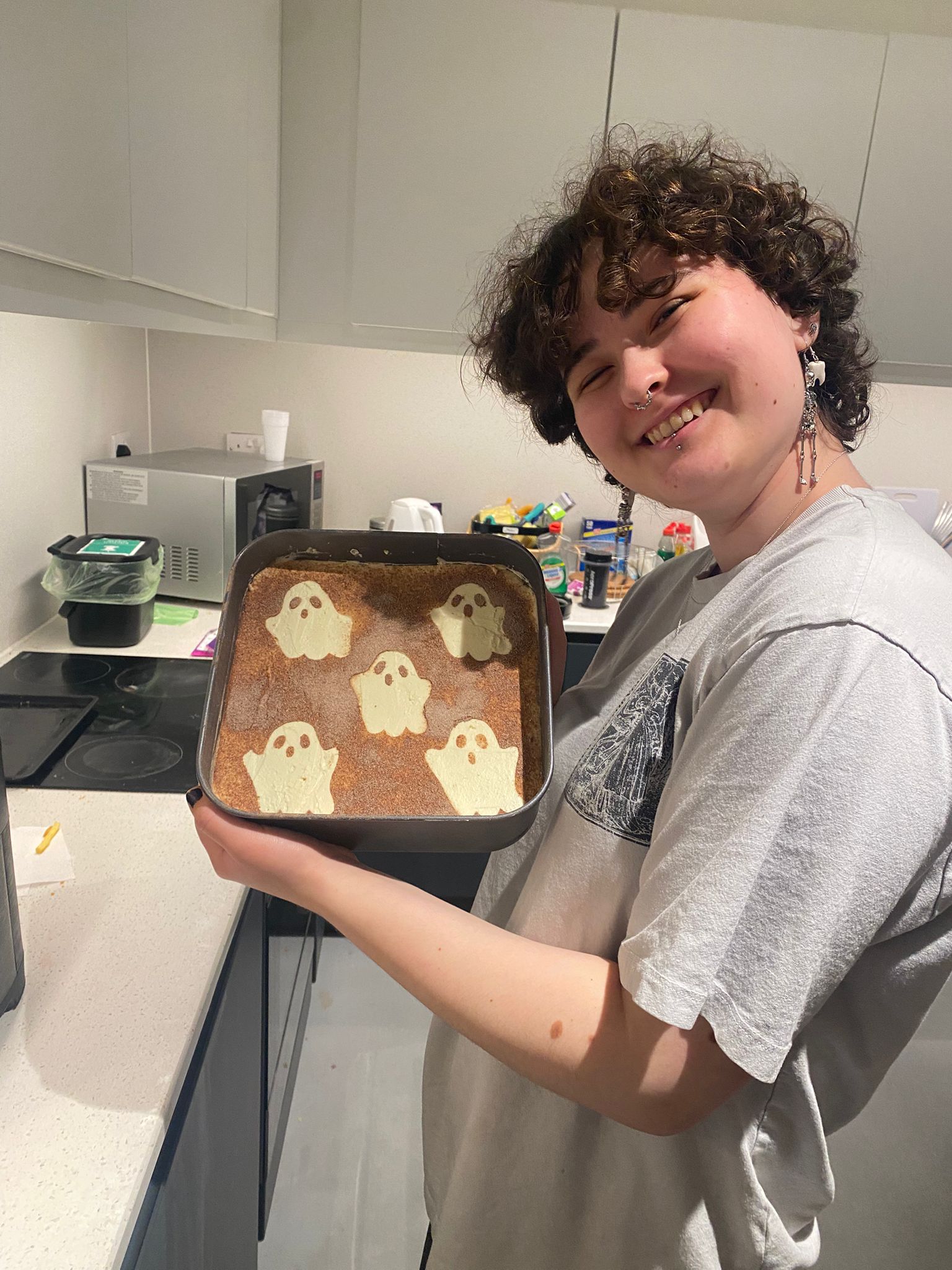 A young person holding a baking tray with a dessert featuring ghost-shaped decorations made from icing. The background includes kitchen appliances and a clean countertop.
