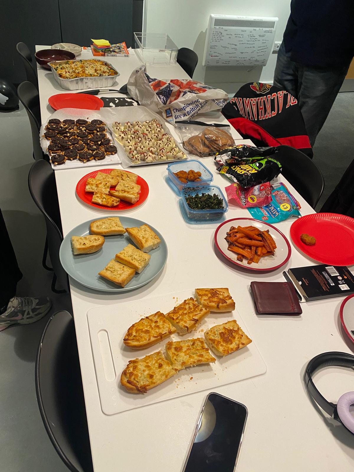 A table covered with various food items including cheese toast, pizza slices, cookies, sweets, and snacks displayed on red and blue plates. Several containers and bags of snacks are scattered across the surface, with a smartphone and headphones visible at the edge.