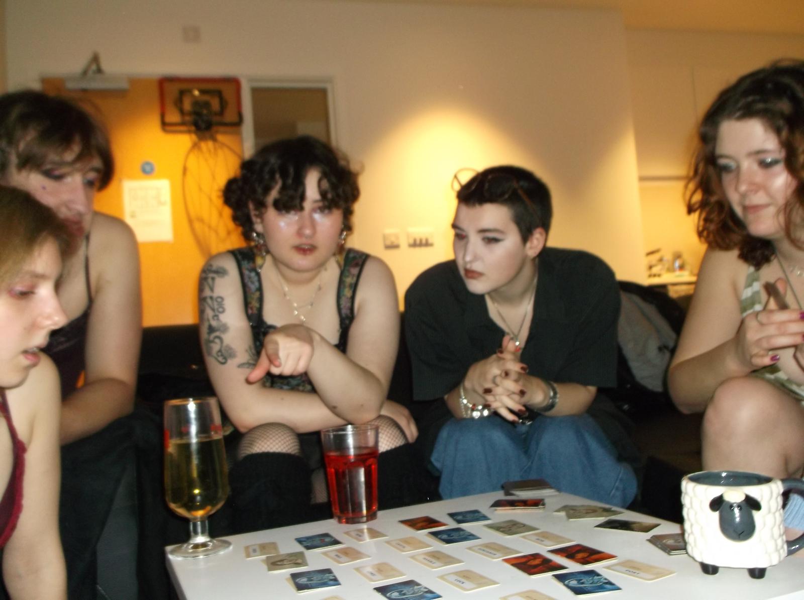 A group of five Arts University Bournemouth students gathered around a table, observing a game with cards laid out on it. Drinks are visible in glasses; the setting is a room with light-coloured walls and a door in the background.
