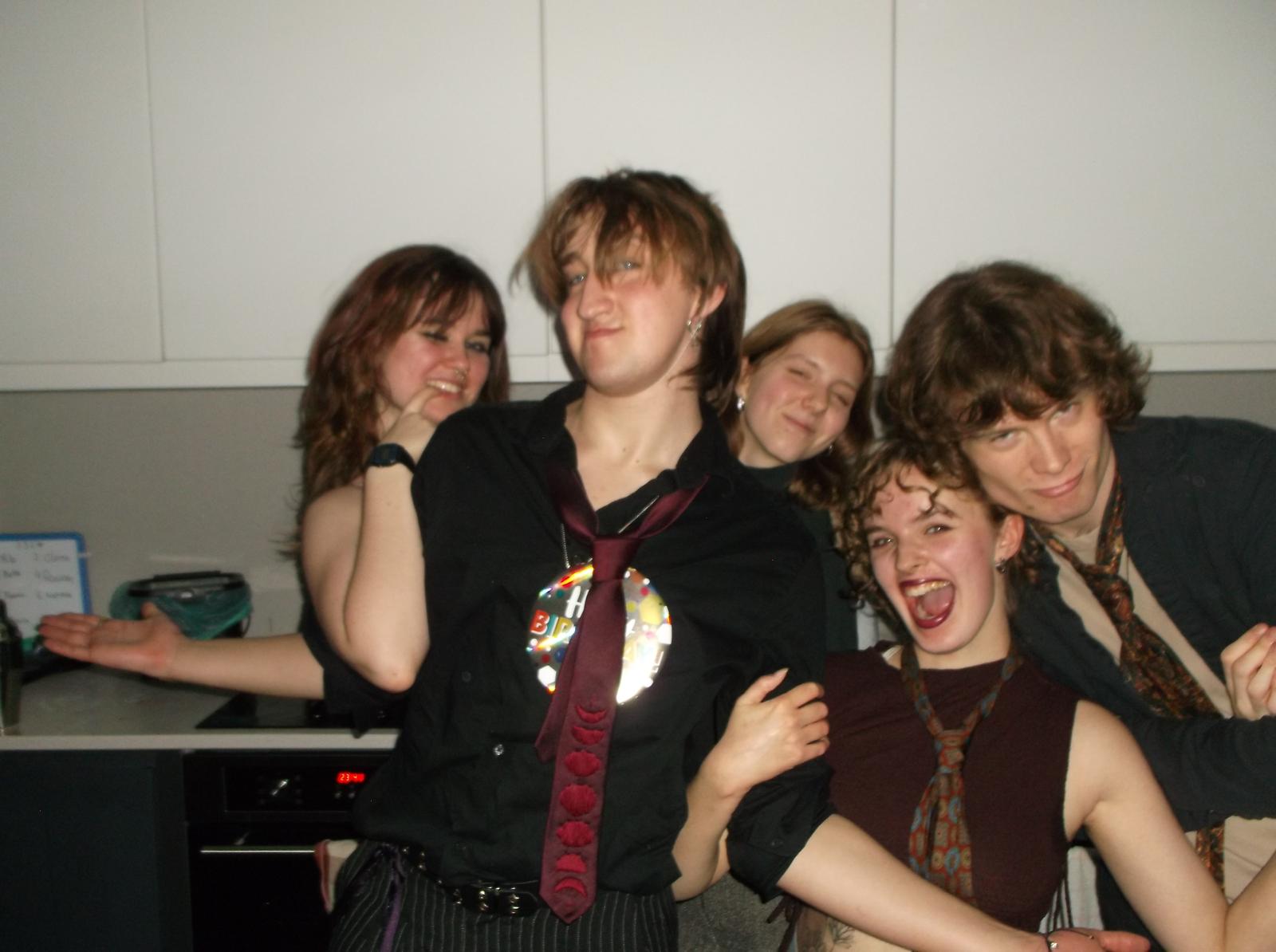 A group of five Arts University Bournemouth students, wearing casual attire, poses playfully together in a kitchen. One person wears a decorative badge, while others have their arms around each other, with a backdrop of white cabinetry and dark appliances.