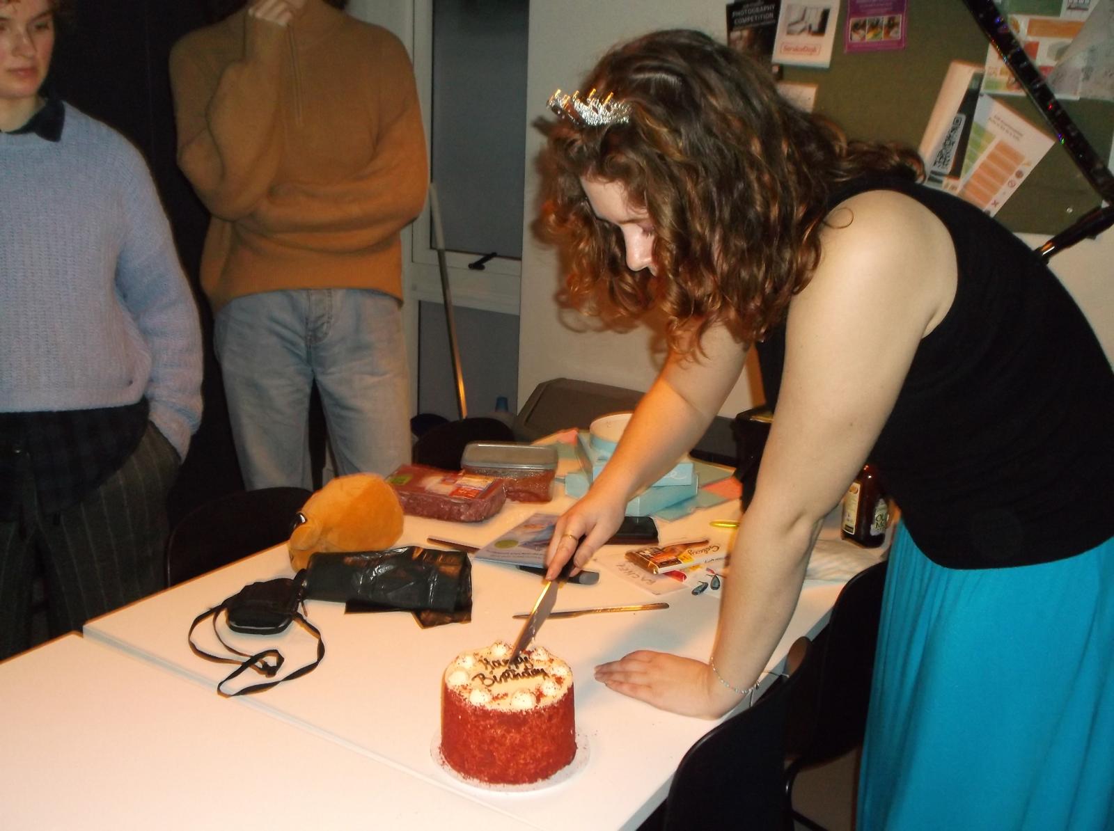 An Arts University Bournemouth student cutting a small cake on a table. Two young people stand in the background, one in a light blue sweater and the other wearing a brown sweater. The setting includes various colourful items and decorations on the wall.