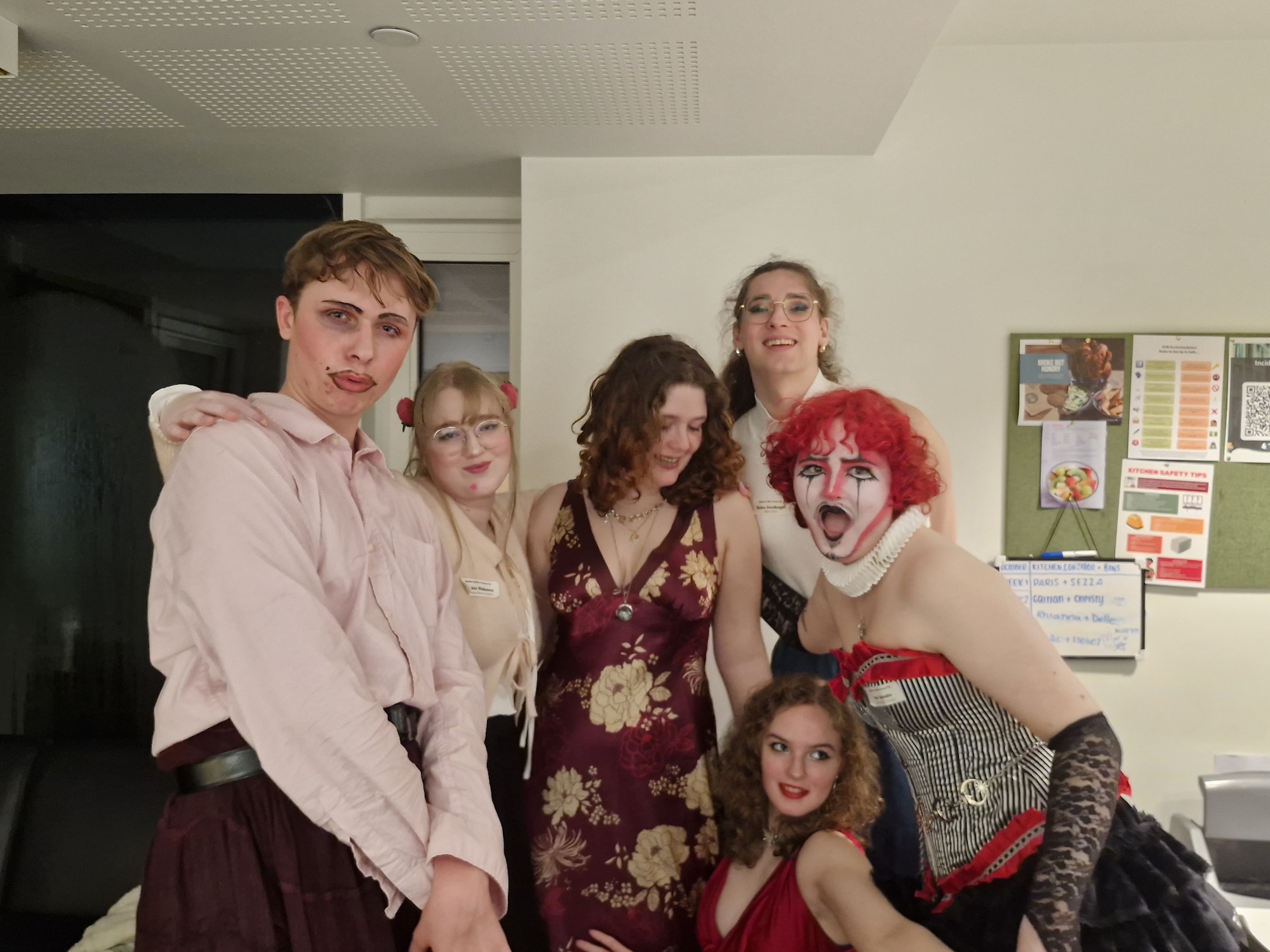 A group of six Arts University Bournemouth students posing together in a brightly lit room. They are dressed in a variety of costumes, with one person wearing clown makeup and outfits ranging from formal to theatrical. A bulletin board with various papers is visible in the background.