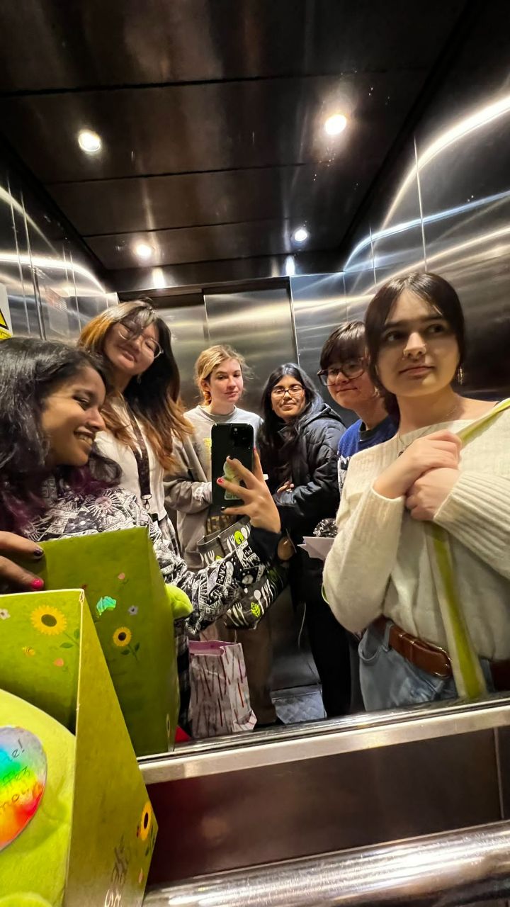 Six people stand close together in a mirrored elevator, with one holding a smartphone for a selfie and colourful shopping bags in the foreground.