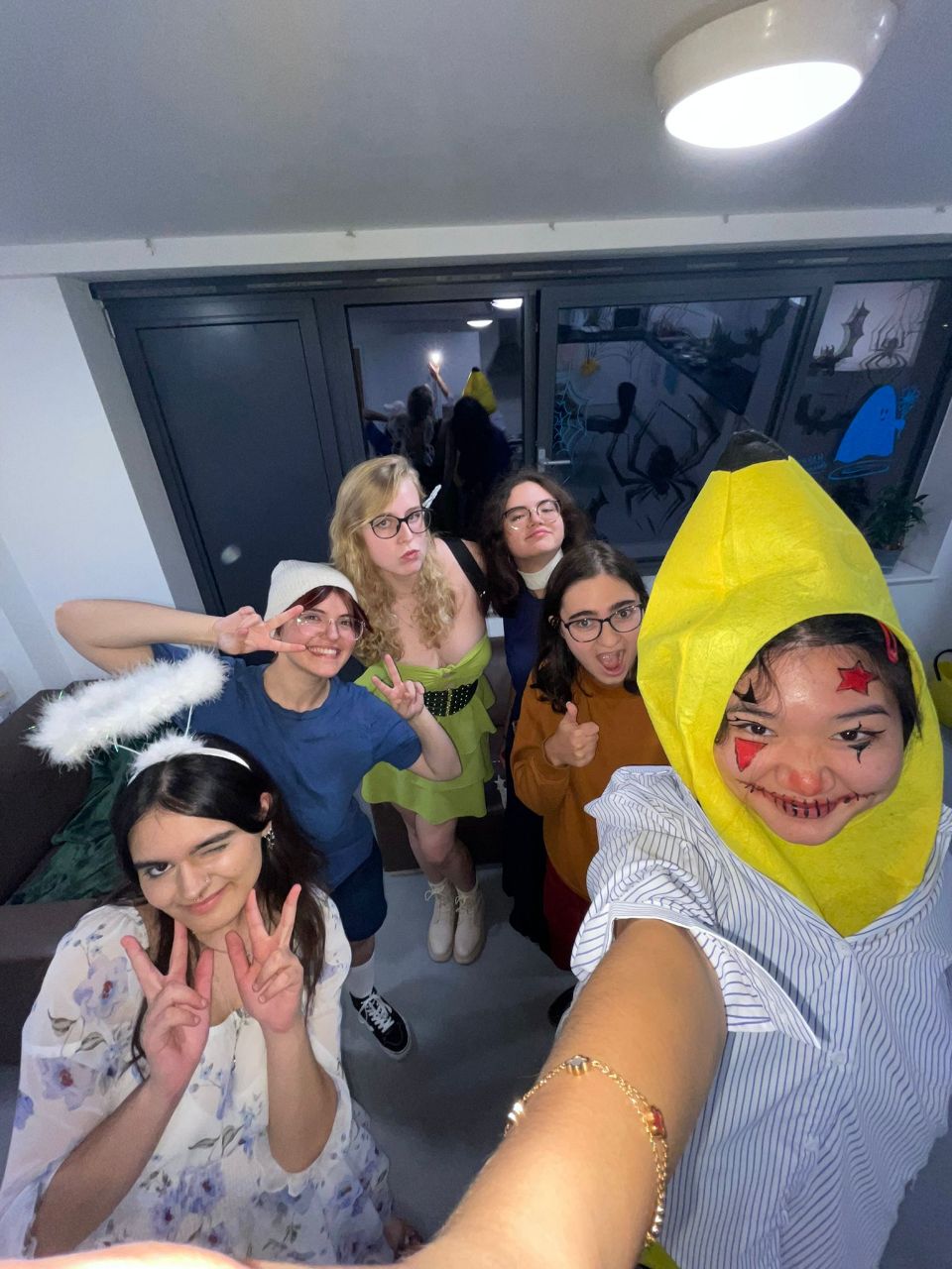 A group of six young adults pose for a selfie indoors, with one person in a bright yellow headpiece and face paint in the foreground.