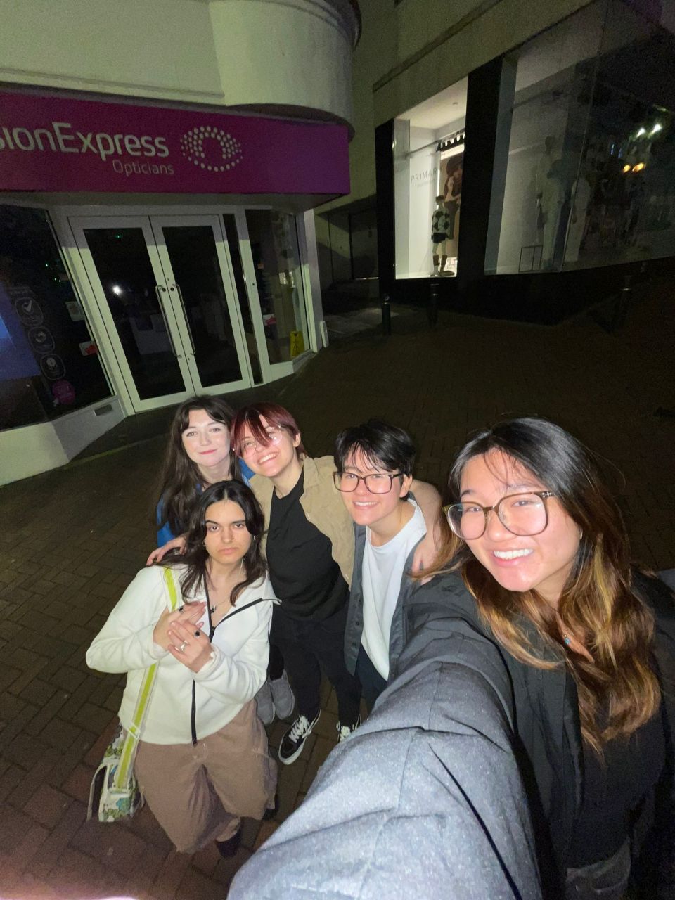 Five young people pose for a selfie outside a Vision Express store at night, with the illuminated shopfront behind them.