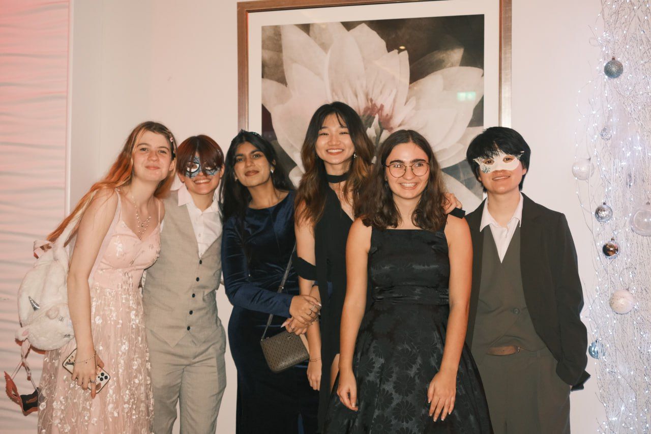 Six Arts University Bournemouth students stand together indoors in formal and semi-formal outfits, with a large flower artwork on the wall behind them.