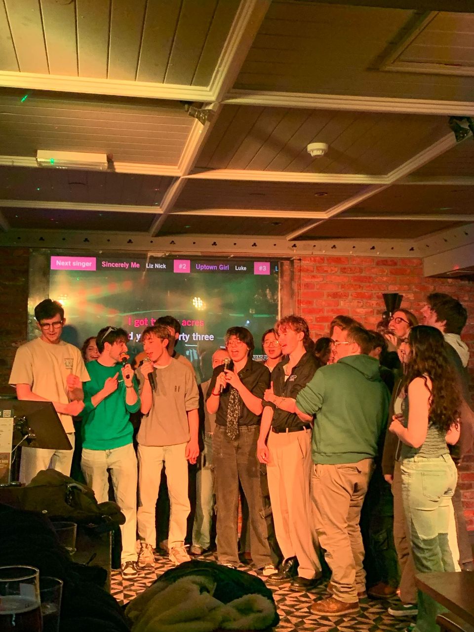 A group of people stands on a small stage in a bar, with a presenter holding a microphone beside a screen and microphone stand.