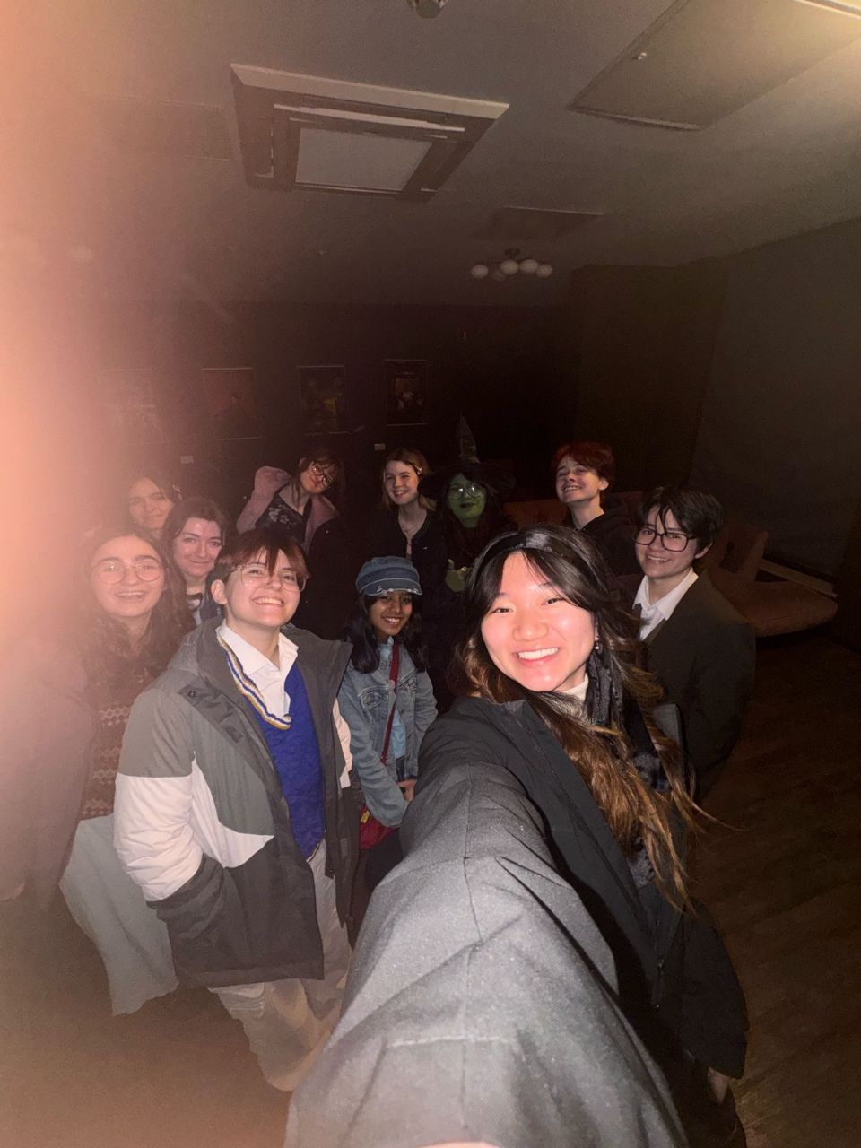 A group of young people posing for a selfie in a dimly lit room, with one person holding the camera in the foreground.