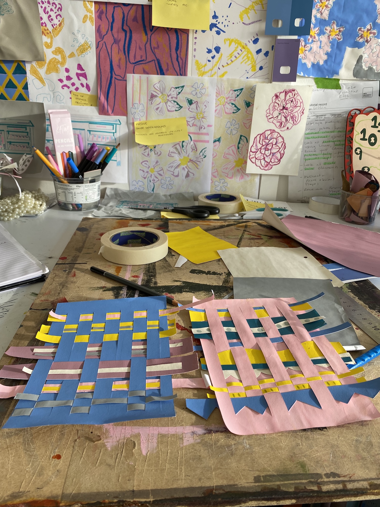 Two colourful paper woven design samples displayed on a studio desk, with art supplies and masking tape surrounding them.