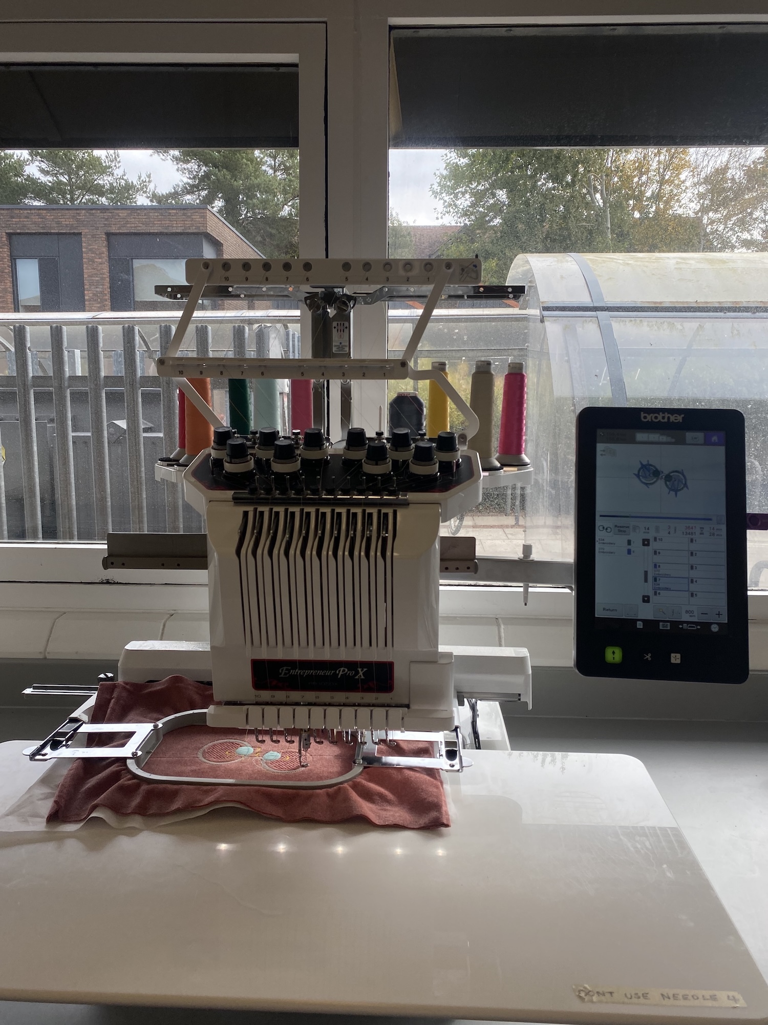 White embroidery machine with multiple coloured thread spools on desk beside black tablet, window showing garden and houses behind.