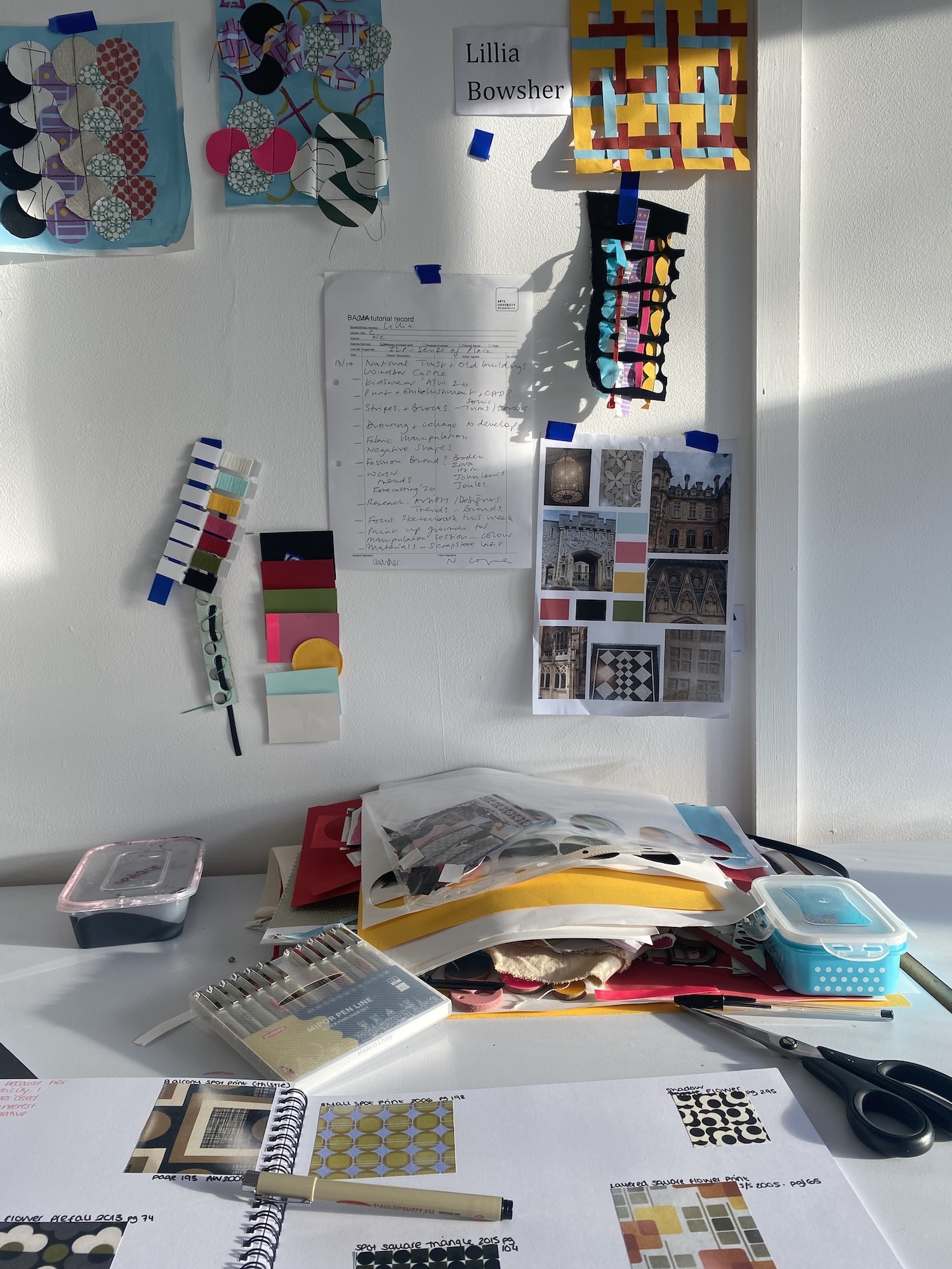 White wall display with colourful artwork, handwritten notes, and material samples. Desk below scattered with design books, colour swatches, and art supplies.