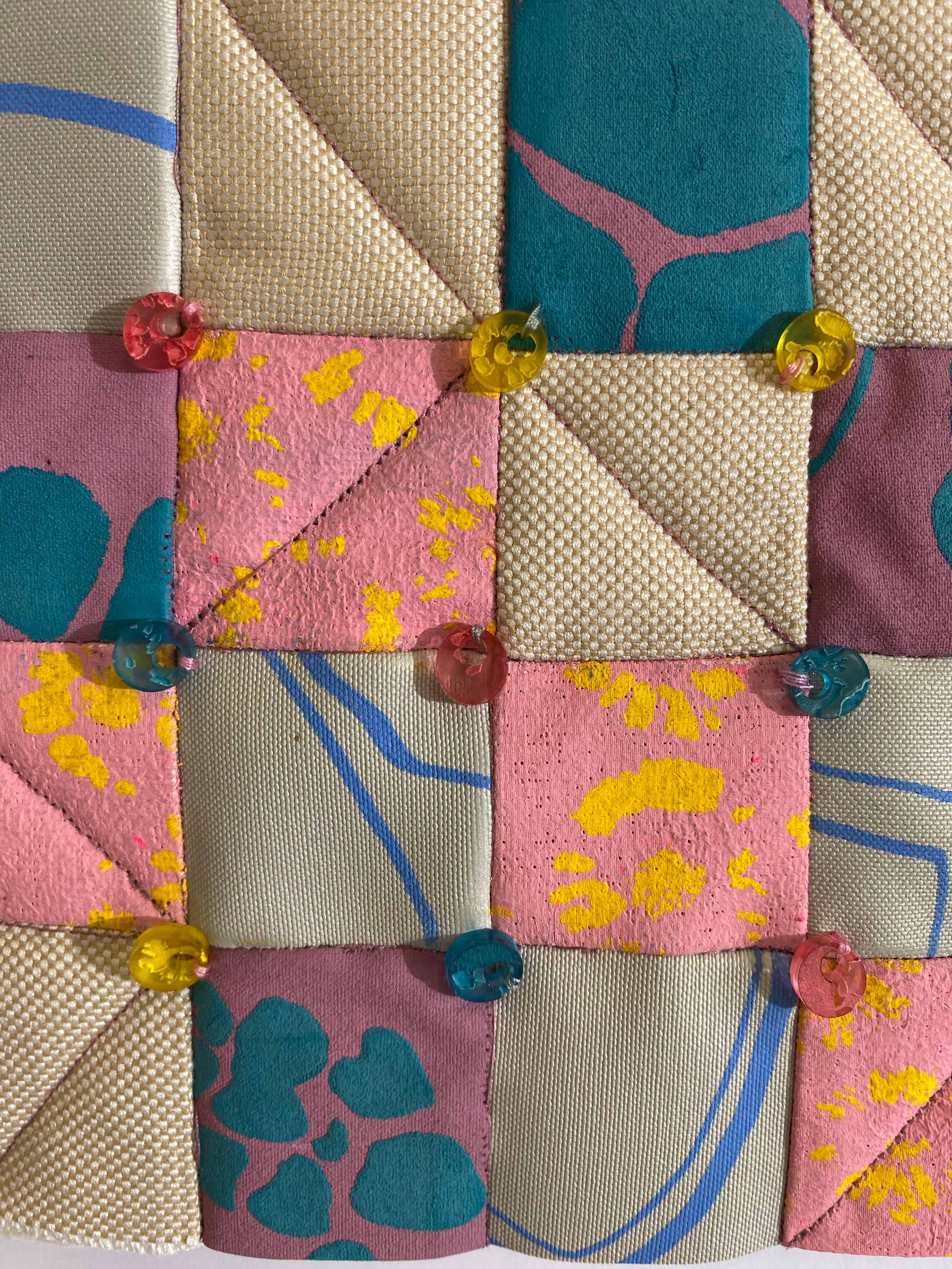 A close-up of a fabric patchwork featuring various textures and patterns, including sections in pink, turquoise, beige, and light grey, with vibrant yellow and pink prints. Small, colourful beads are sewn at the intersections of the patches.