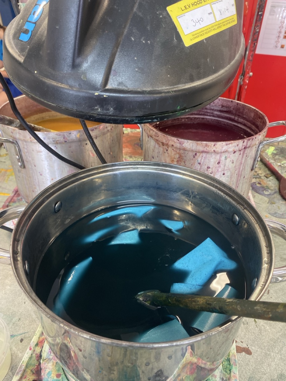 A large metal pot containing blue liquid is shown with a stirring stick inside. In the background, two other pots with yellow and burgundy liquids are visible. The scene is set on a surface with splashes of paint.