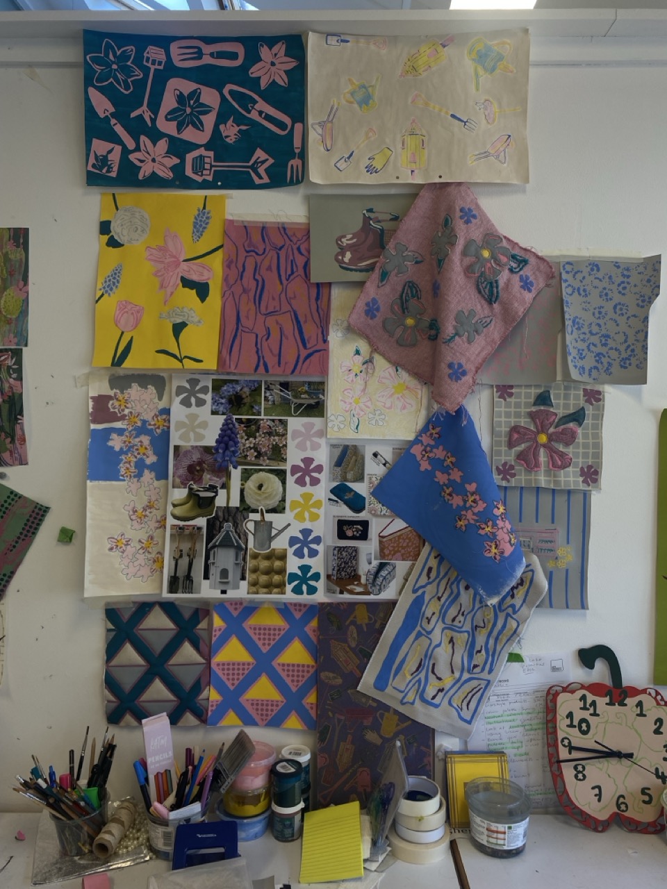A collection of artworks displayed on a wall, featuring various floral and abstract patterns in bright colours. Below, there are art supplies including pens, paint cans, and a yellow notepad, along with a large clock in the corner.