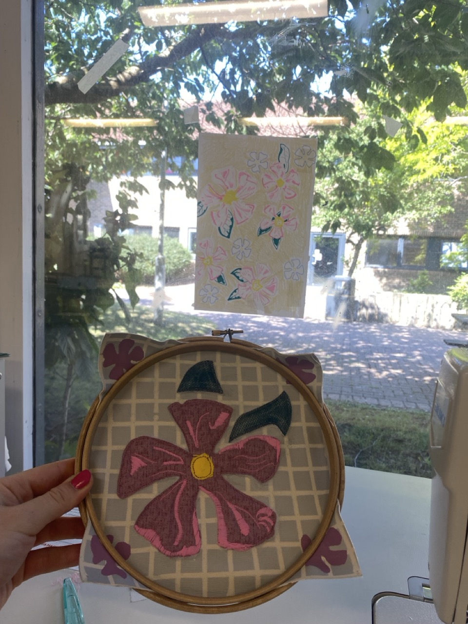 A person holds a circular embroidery hoop featuring a large pink flower with yellow centre against a patterned background. In the background, a drawing of flowers is taped to a window, revealing greenery outside.