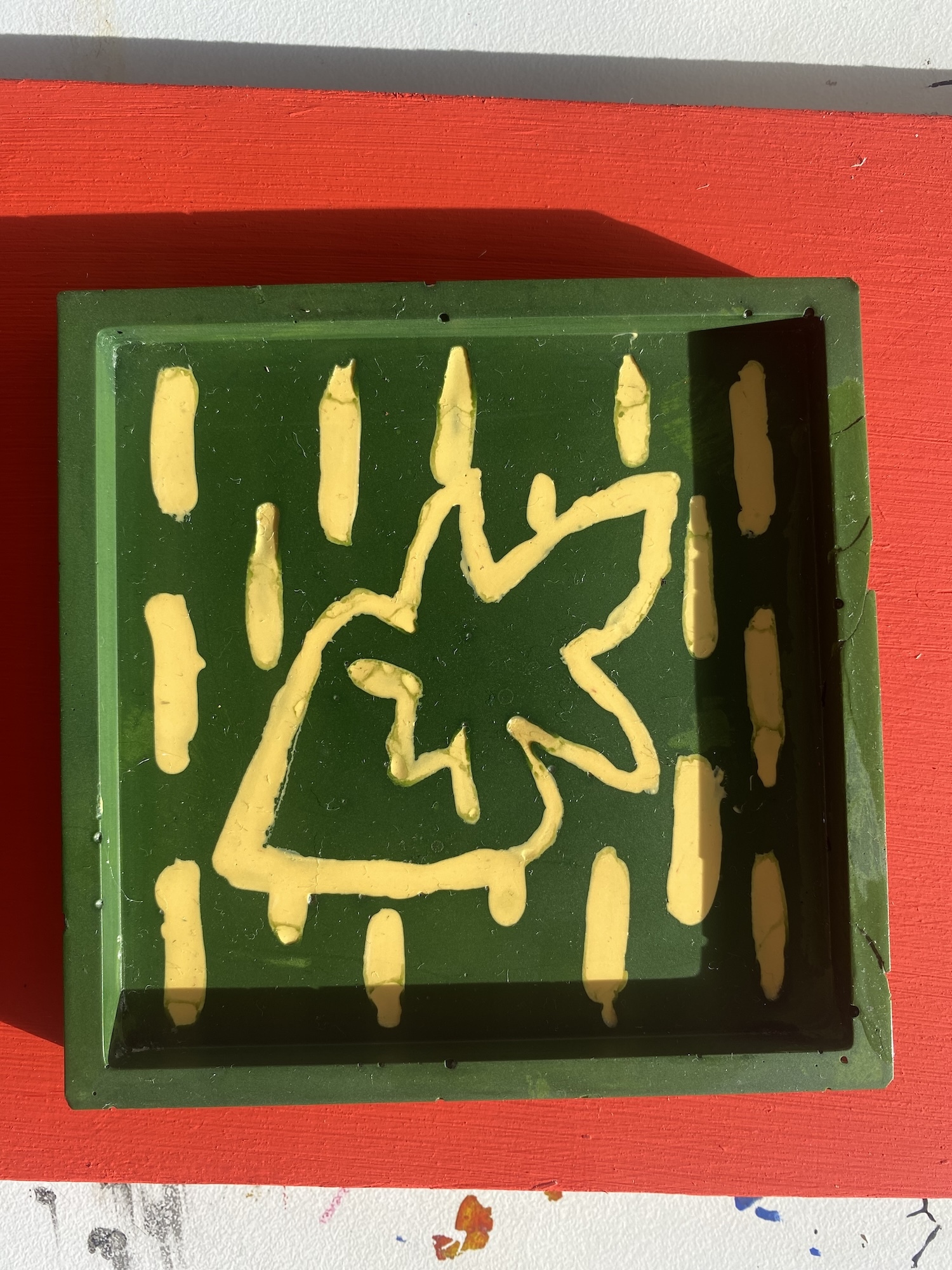 Green glazed Jesmonite tile with yellow paintwork, placed on bright red surface with white edges visible.