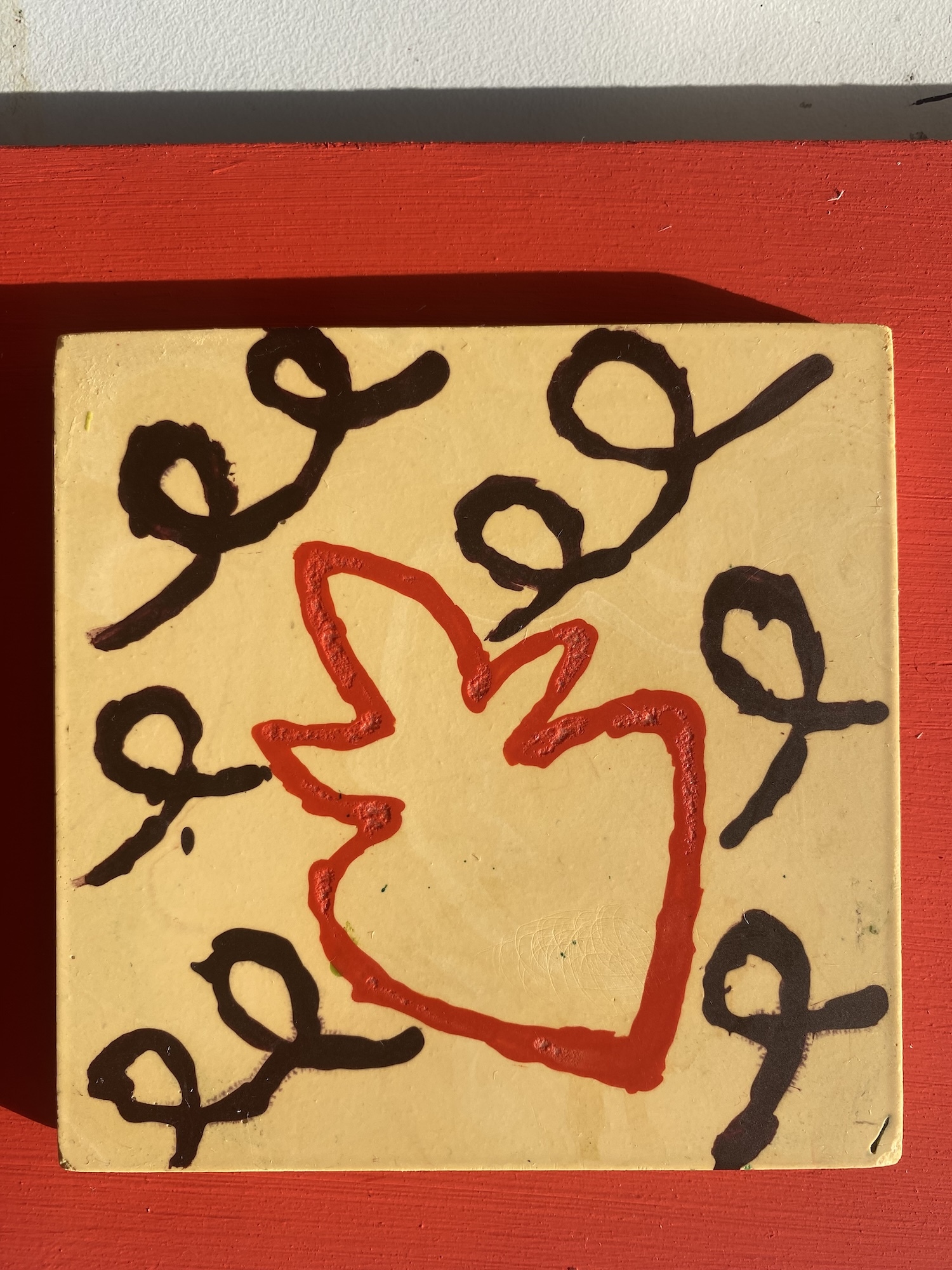 Yellow Jesmonite square tile with red heart outline in centre, surrounded by black curved lines with loops, mounted on red surface.