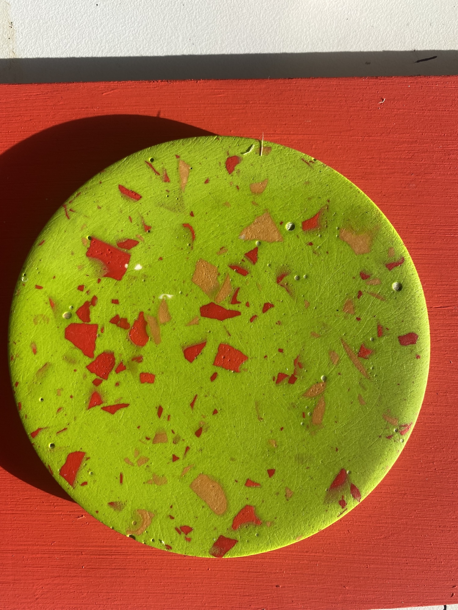 Bright green circular Jesmonite tile with red and orange speckles scattered across surface, placed on red wooden background.