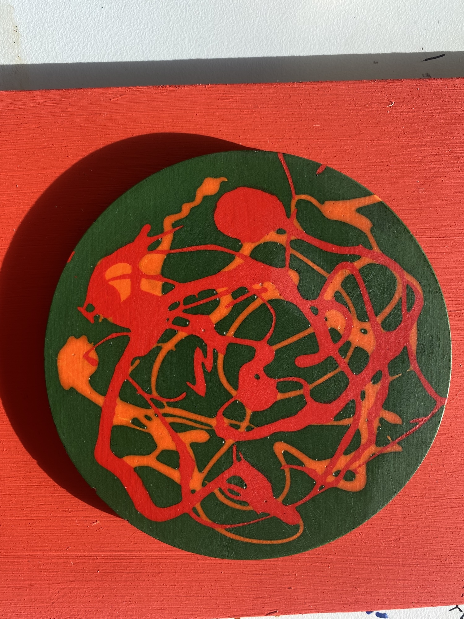 Circular green Jesmonite tile with red and orange abstract linear patterns inside, painted on bright red background with grey stripe above.