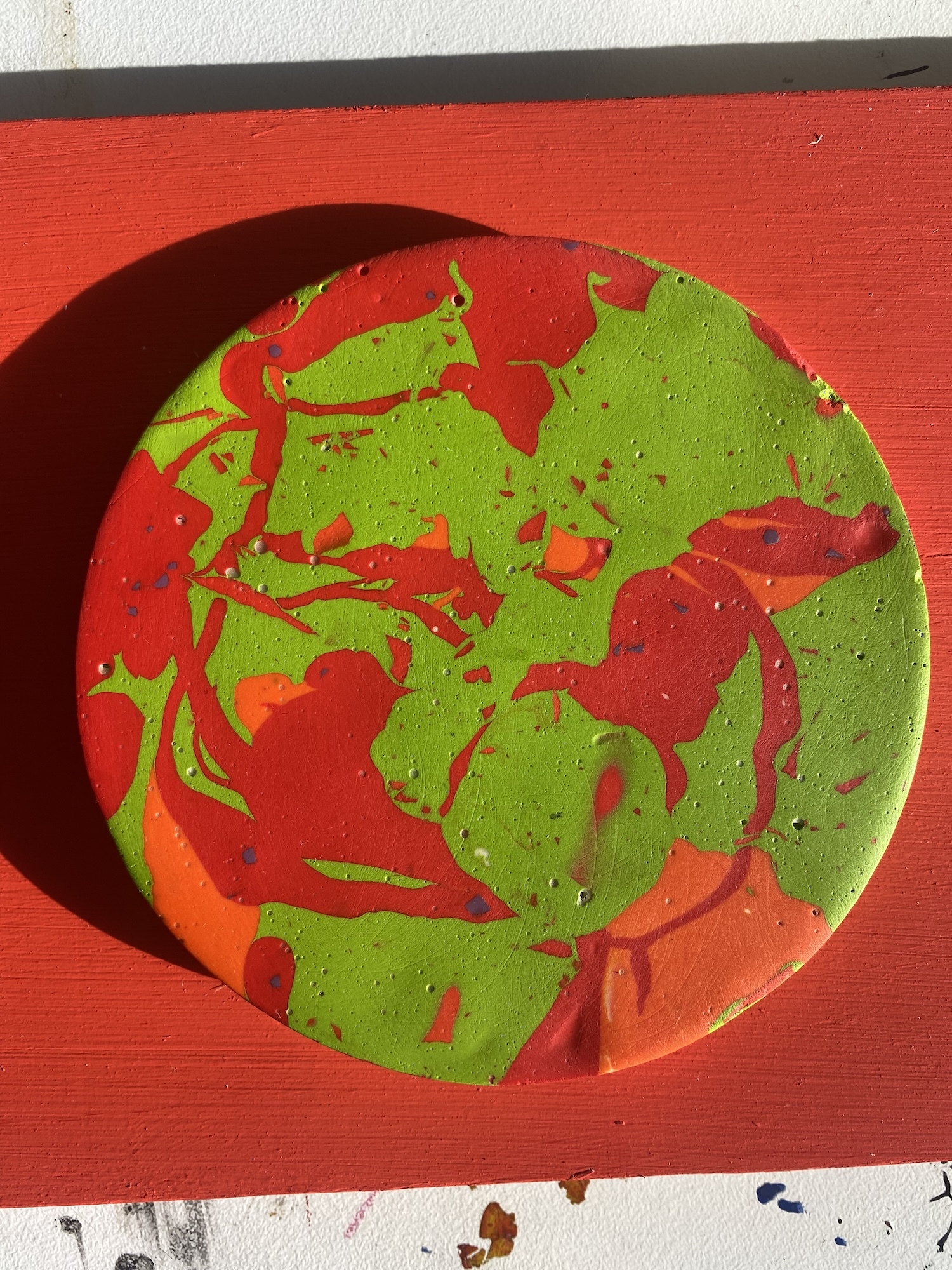 Circular painting with swirled red and bright green paint on red Jesmonite surface, creating marbled pattern within round shape.