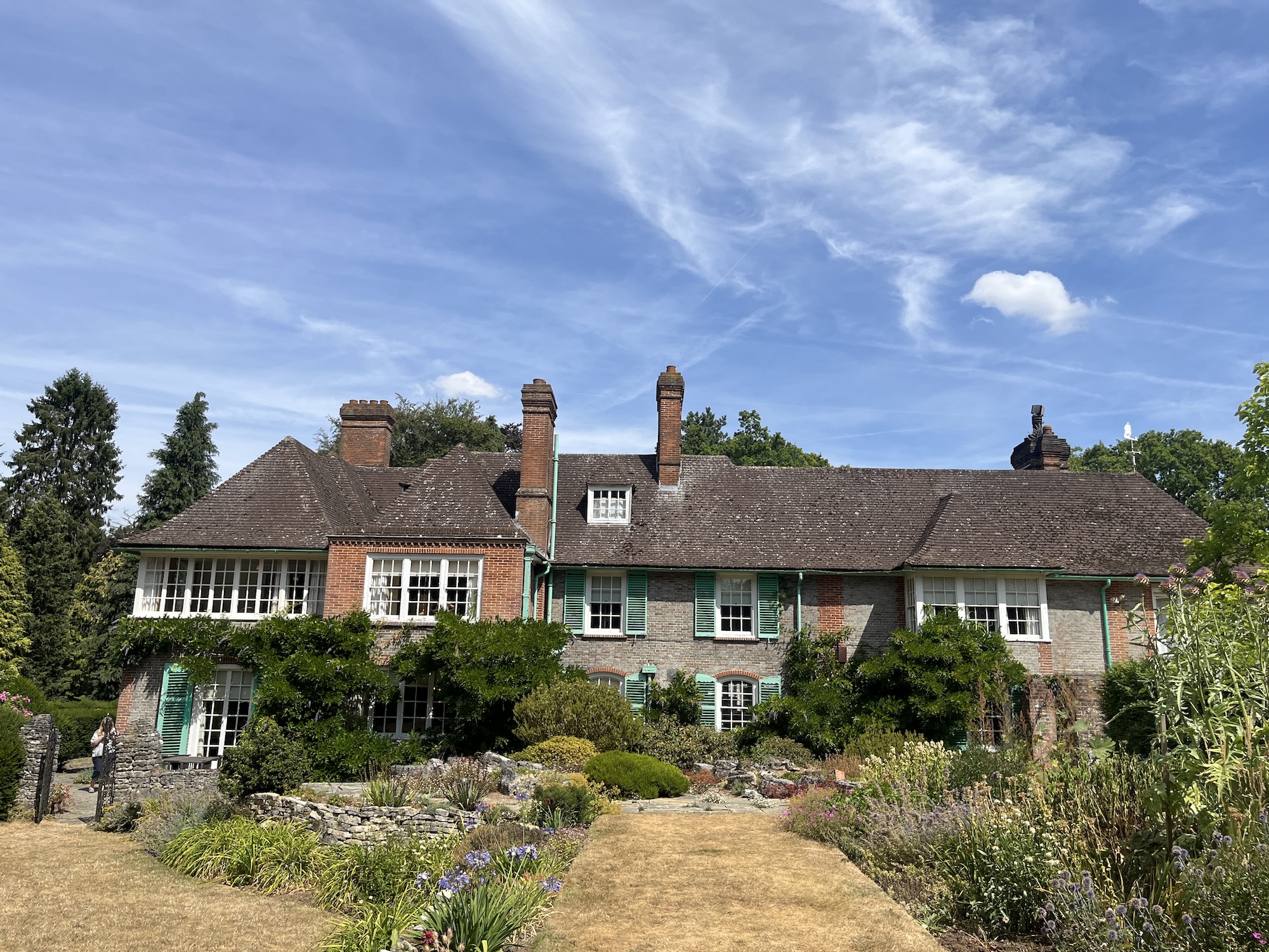 A long brick and timber country house with chimneys and green-trimmed windows stands behind a flower-filled garden and dry lawn under a blue sky.