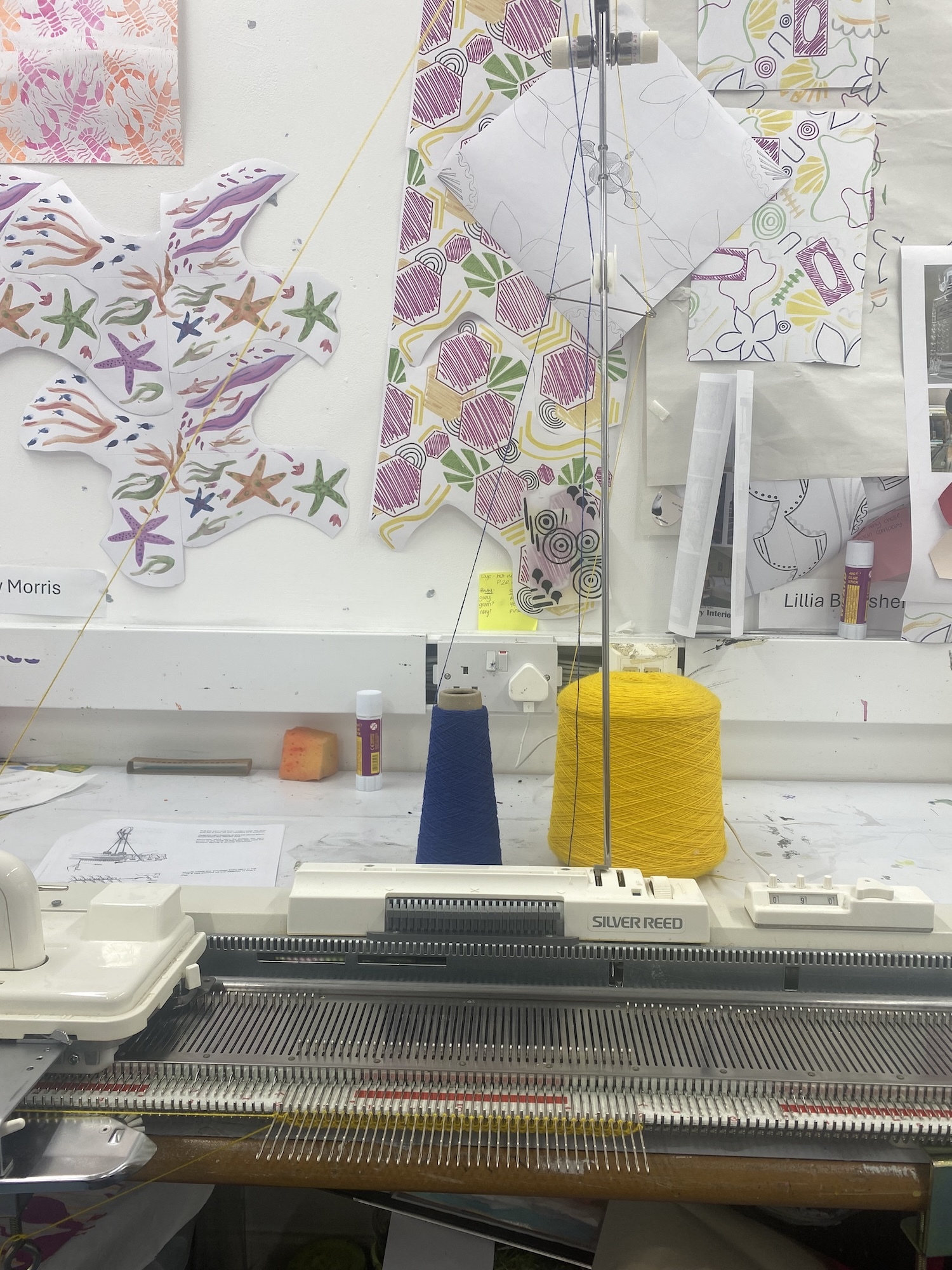 Knitting machine with yellow yarn and blue cone weight. Colourful pattern samples pinned to white wall above workspace.