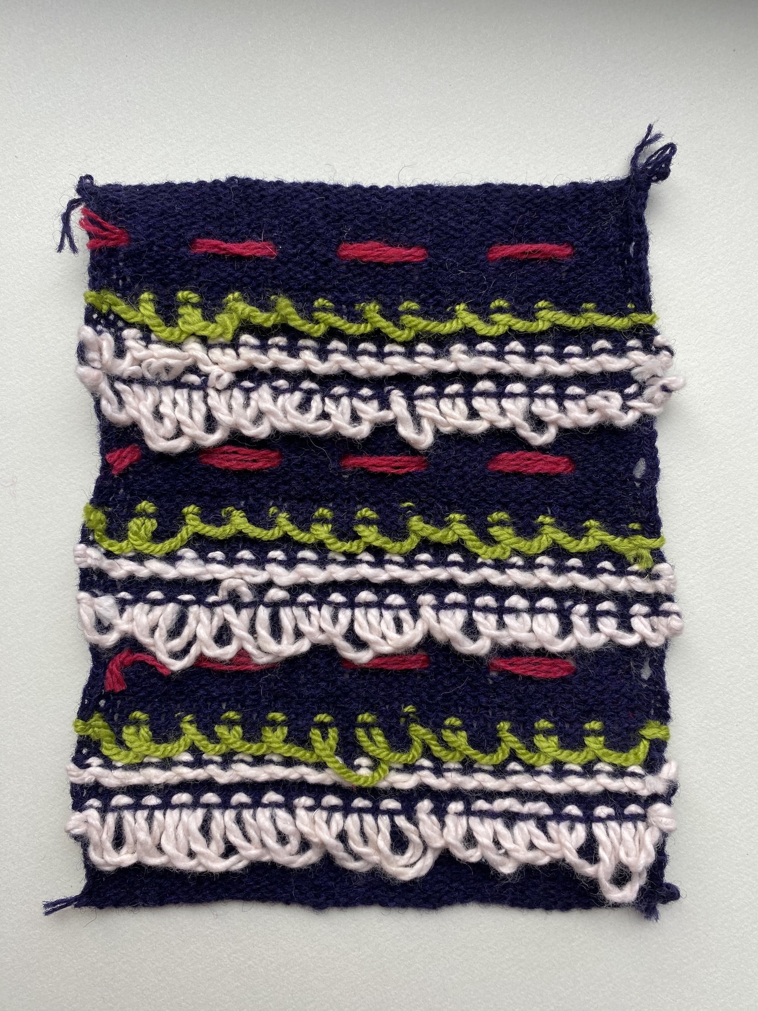 Knitted textile sample with navy blue base, red dashes, lime green zigzag trim, and white looped fringe in horizontal bands.