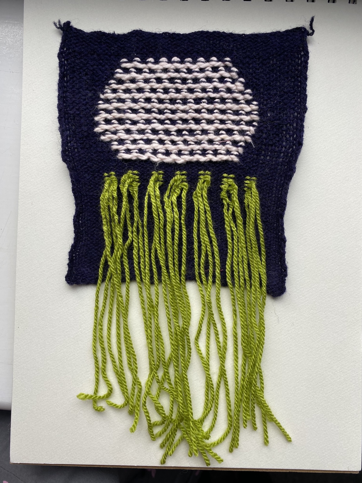 Navy blue knitted wall hanging with white circular pattern in centre and long green yarn tassels hanging below.