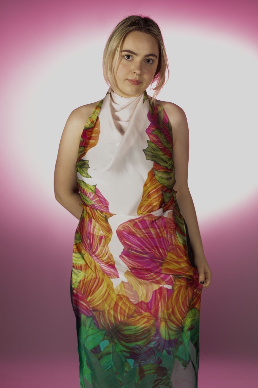 Woman wearing white halter top and tropical print sarong with pink, orange, yellow and green leaf pattern against pink gradient background.