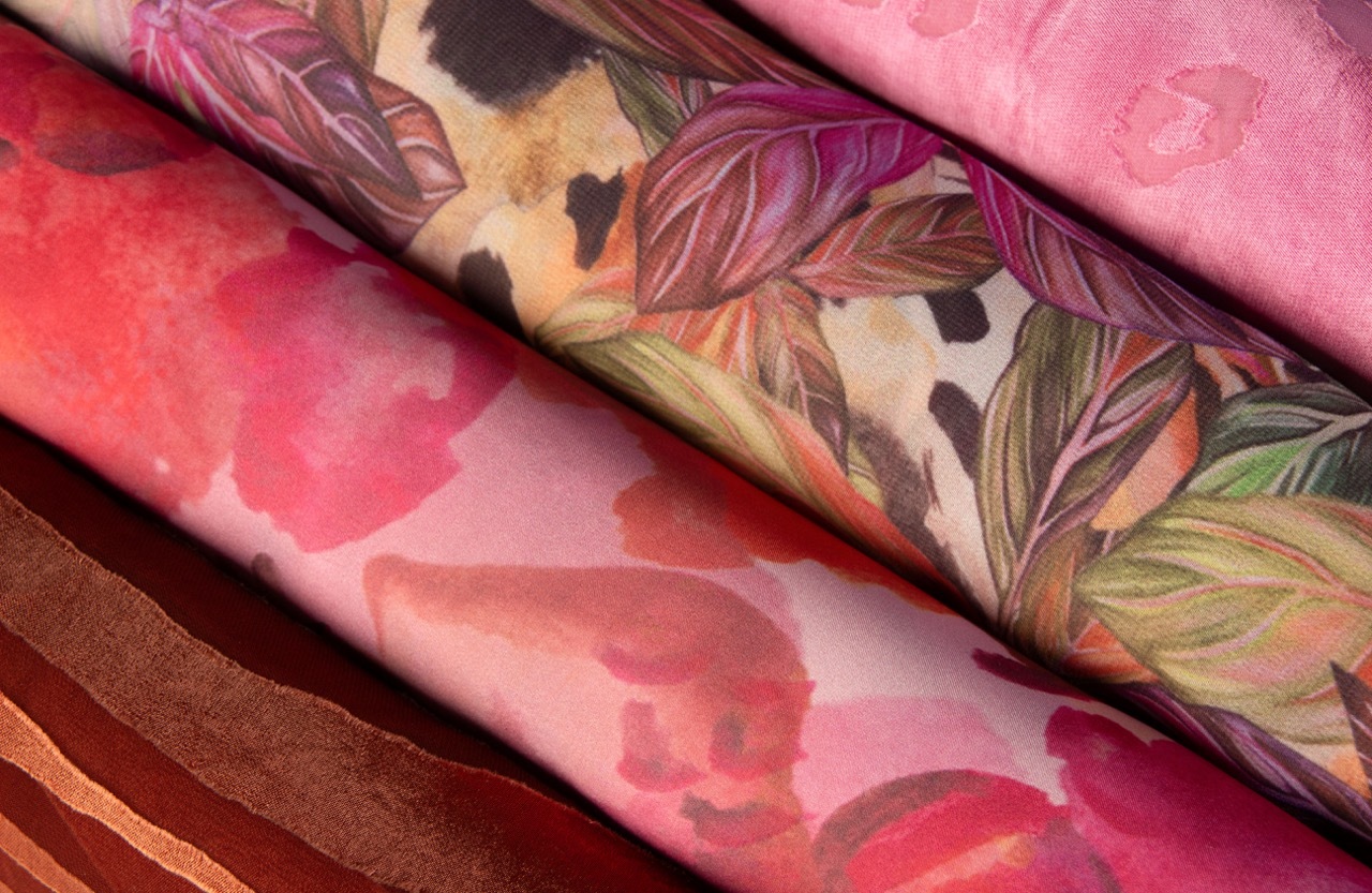 Three folded fabric pieces with floral patterns in pink, burgundy, and cream tones arranged diagonally across wooden surface.
