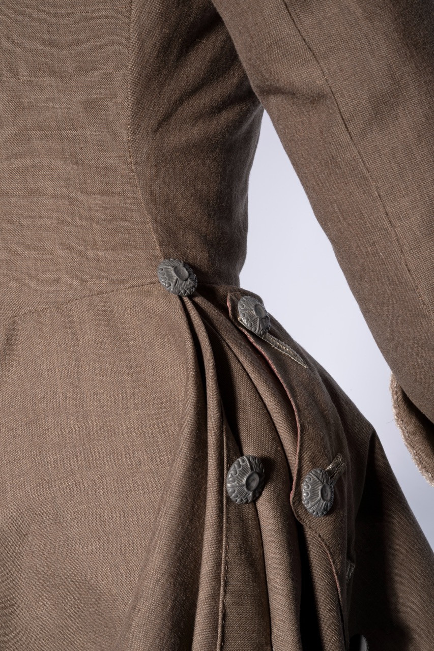 A close-up view of a brown garment featuring intricate pleats and decorative buttons. The fabric shows texture, highlighting the garment's tailored design and craftsmanship, with the background being a neutral white.
