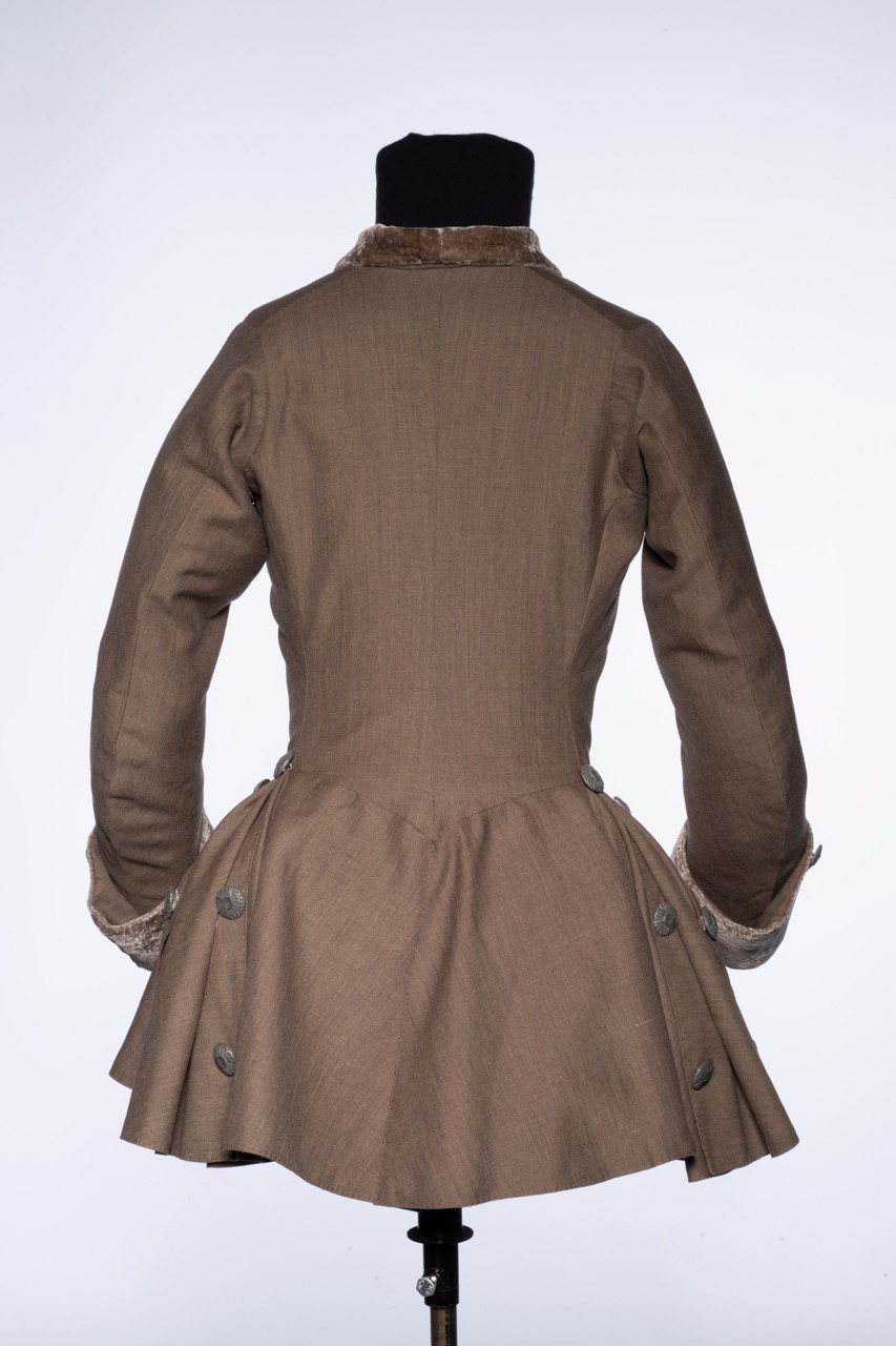 A brown linen jacket displayed on a mannequin, showing the back view. The jacket features a fitted bodice, a flared skirt, and is accented with silver buttons along the back and a fur collar.