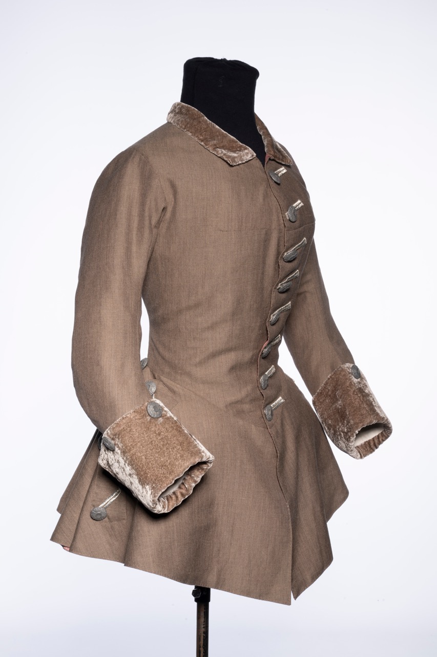 A historical brown coat displayed on a black mannequin, featuring a high collar and velvet cuffs. The coat has a fitted bodice that flares out at the waist, equipped with decorative buttons down the front.