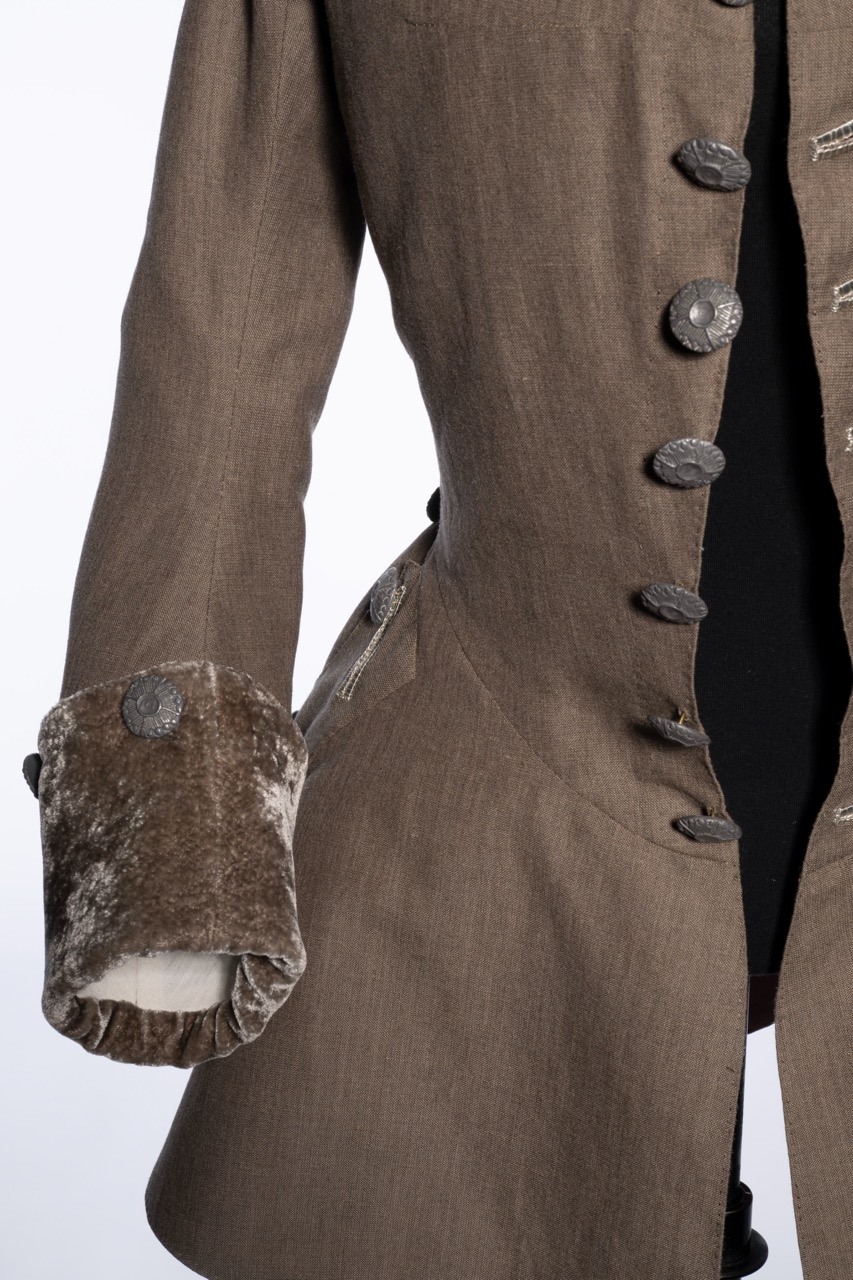 A close-up of the sleeve and front buttons of a brown tailored coat. The sleeve features a velvet cuff with a button, while the coat has a textured finish and multiple buttons down the front.