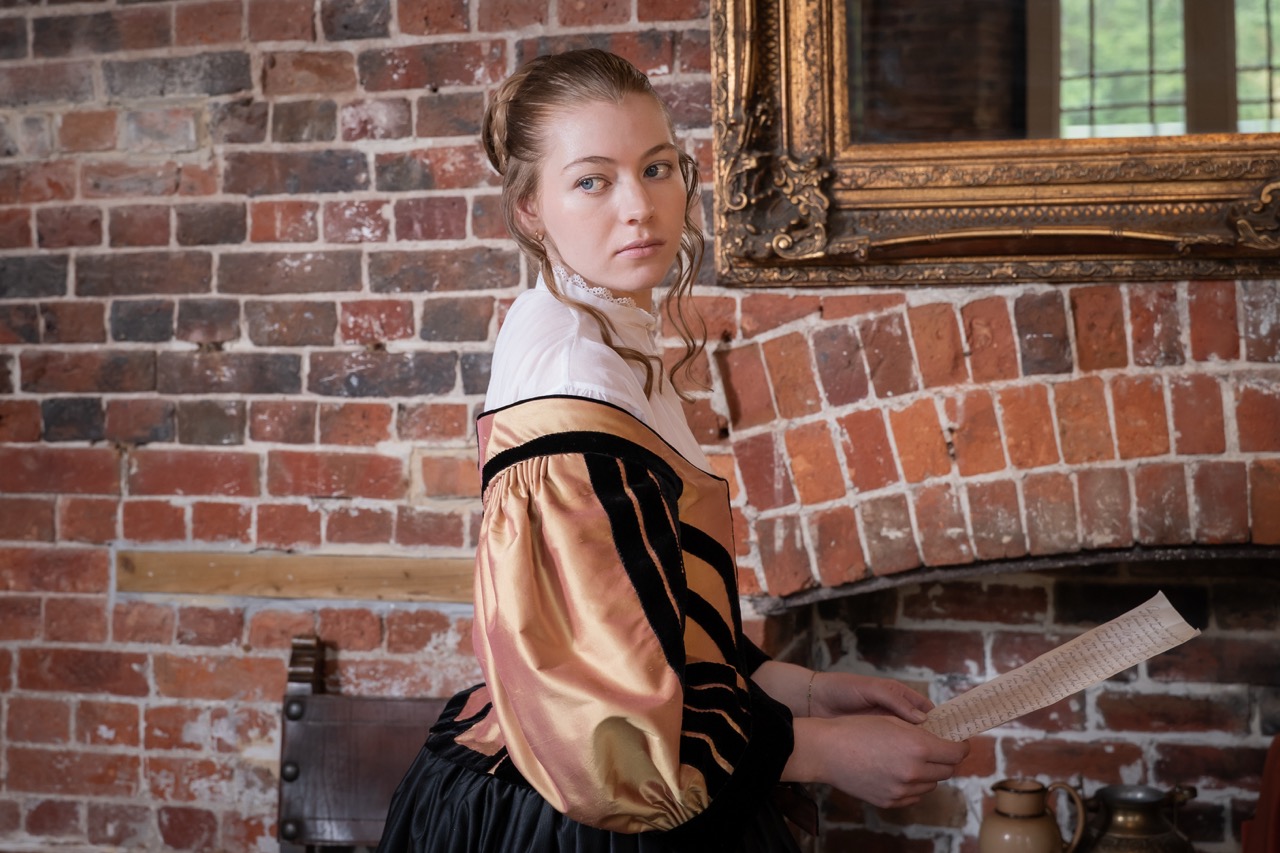 A young woman with long hair wearing a historical dress with puffed sleeves stands near a rustic brick fireplace. She holds a piece of paper in one hand, and a gilded mirror is visible on the wall behind her.