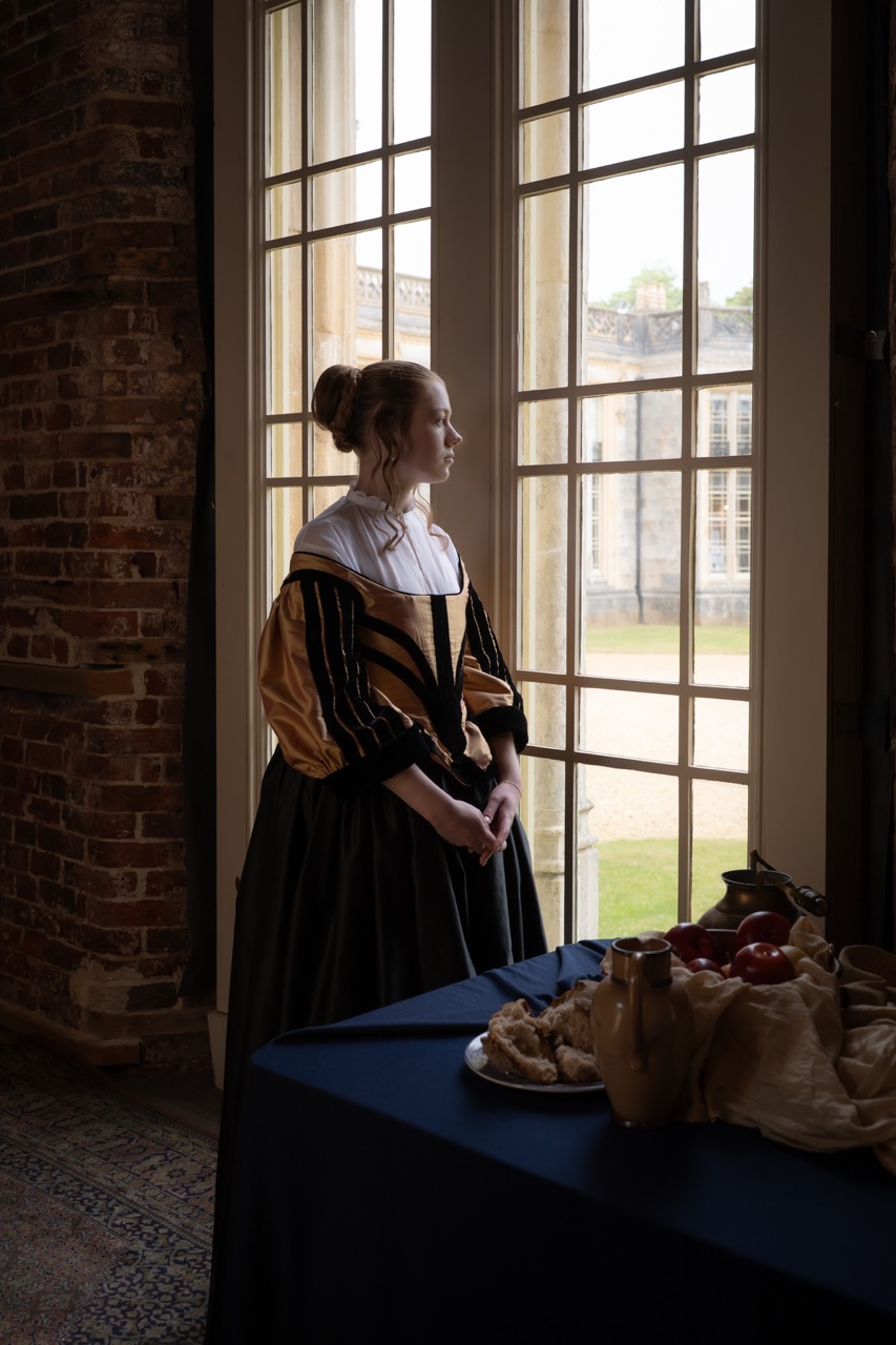 A woman wearing a historical dress with a black and orange bodice stands by large windows, looking outside. A table with a cloth features various items, including a bowl and a basket in a room with brick walls and natural light.