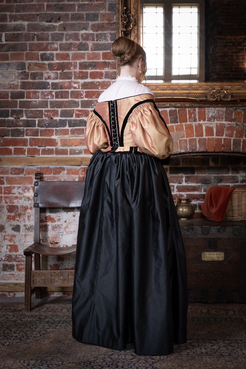 A woman is viewed from behind, wearing a historical dress with a fitted bodice and a voluminous black skirt. The dress features a peach-coloured blouse with puffed sleeves and a white collar. The background includes exposed brick walls and a wooden chair.