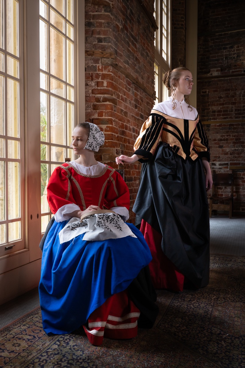 Two women in historical costumes with elaborate details stand in a room featuring brick walls and large windows. One woman, seated, wears a red and blue dress with a white bonnet, while the other stands beside her in a black and gold gown.
