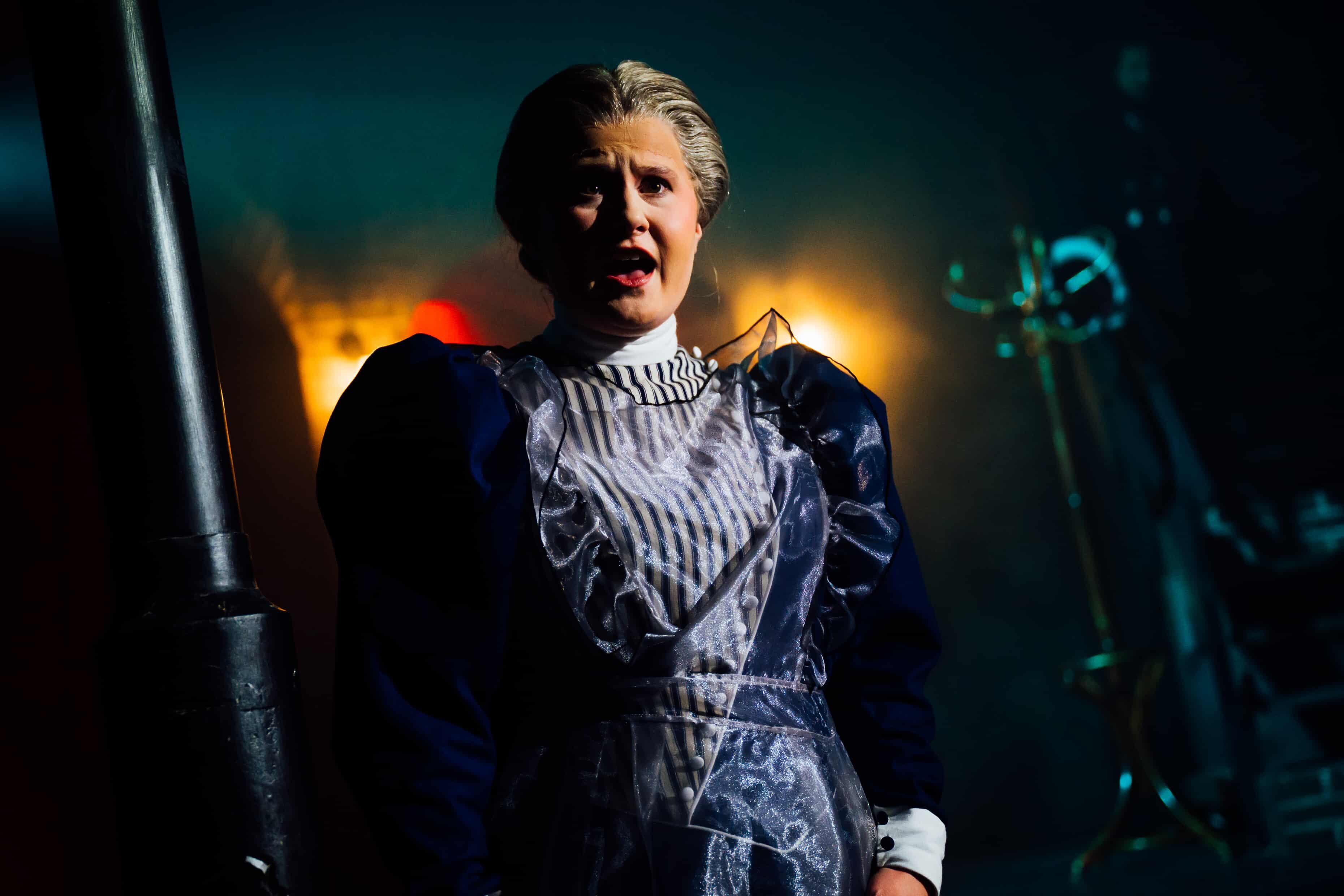 A person wearing an elaborate dark dress with puffed sleeves stands in a dimly lit environment. The background features soft glowing lights and a clothing rack, while the figure is illuminated from the front.