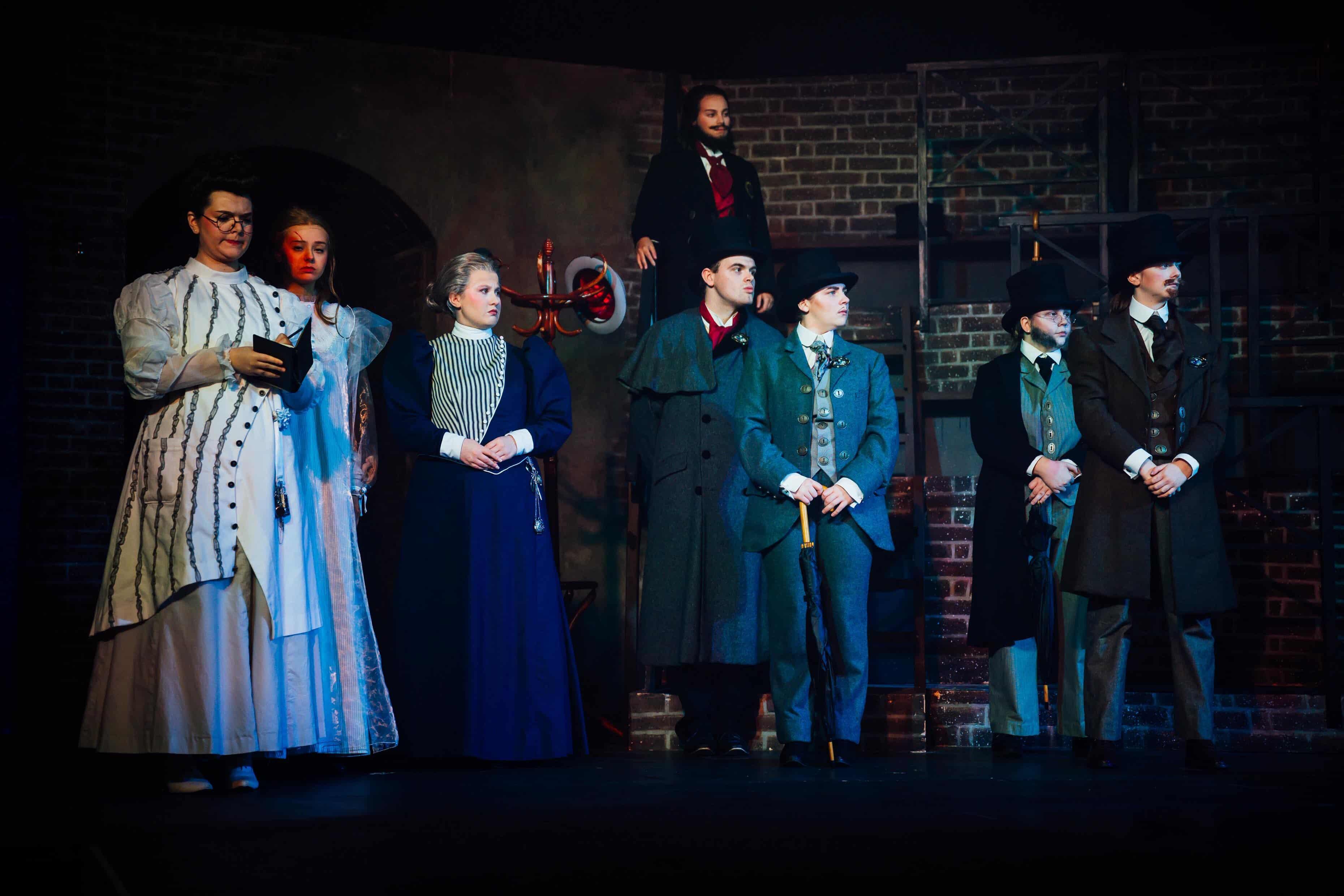 A group of six actors dressed in period costumes stands on a stage with a brick wall backdrop. They include a woman in a long white dress with a black corset, a woman in a blue dress, and men in dark suits, some holding canes.