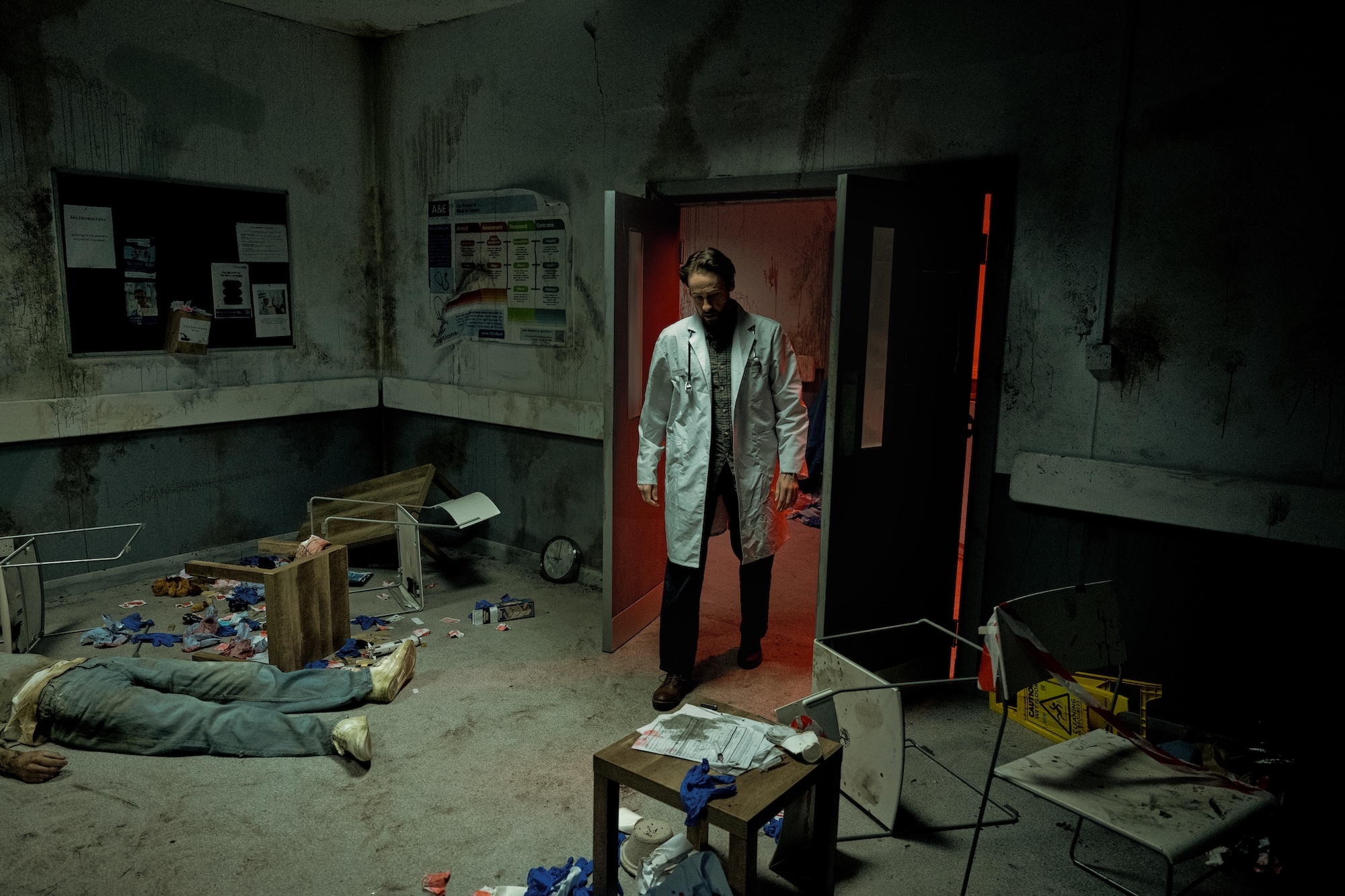 Man in white coat standing in doorway of dimly lit, cluttered room with scattered papers, boxes, and debris on floor.